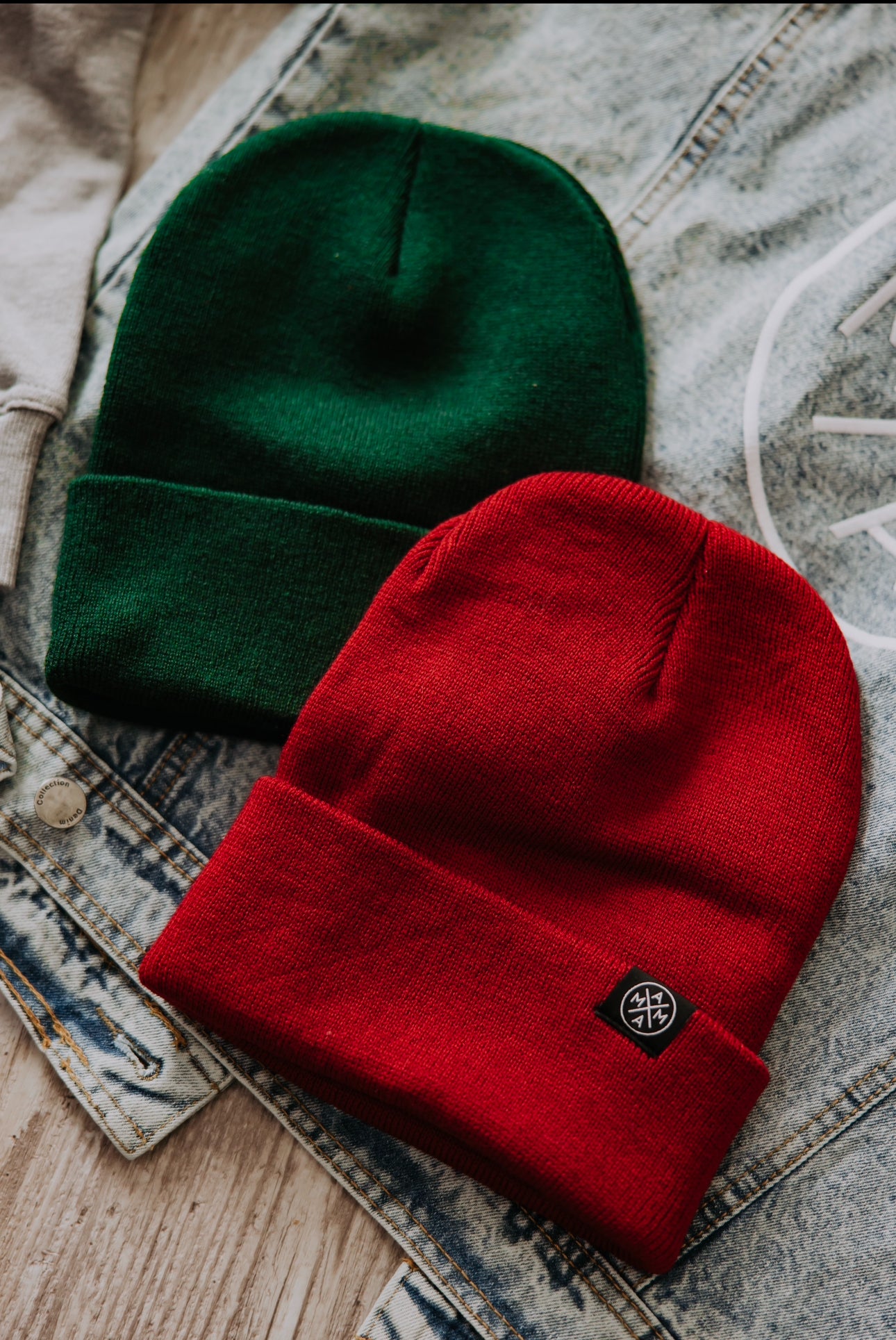 Two Mama X™ cuffed beanies are placed on a denim jacket. One 12-inch cuffed beanie is green, while the other is a red Mama X™ creation featuring a small logo patch on the cuff. Both are laid out on a wooden surface.