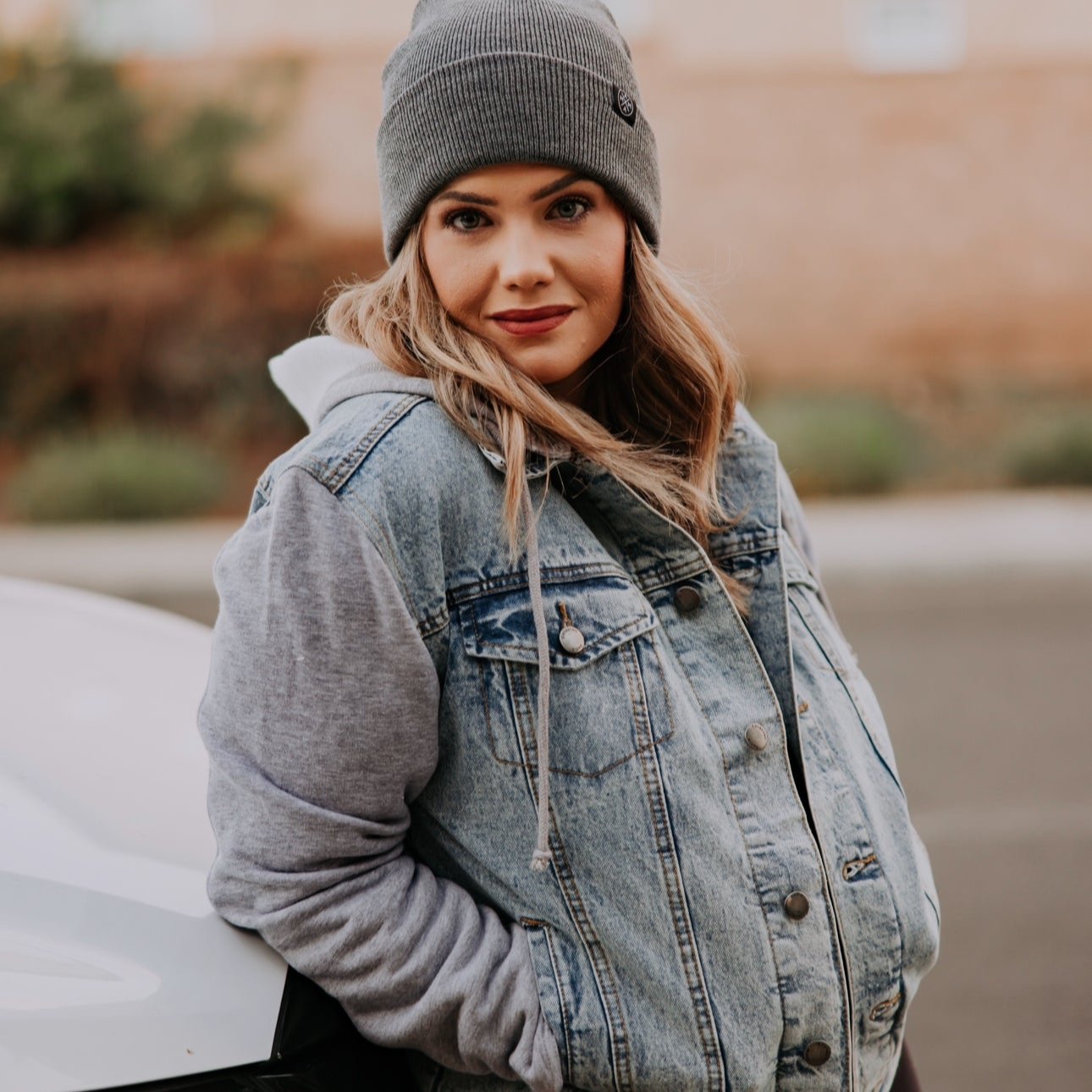 A person clad in a Mama X™ Embroidered Jean Jacket, layered over a hoodie, wears a gray beanie and dark pants while leaning casually against a white car. The backdrop includes a blurred brick wall complemented by some greenery.