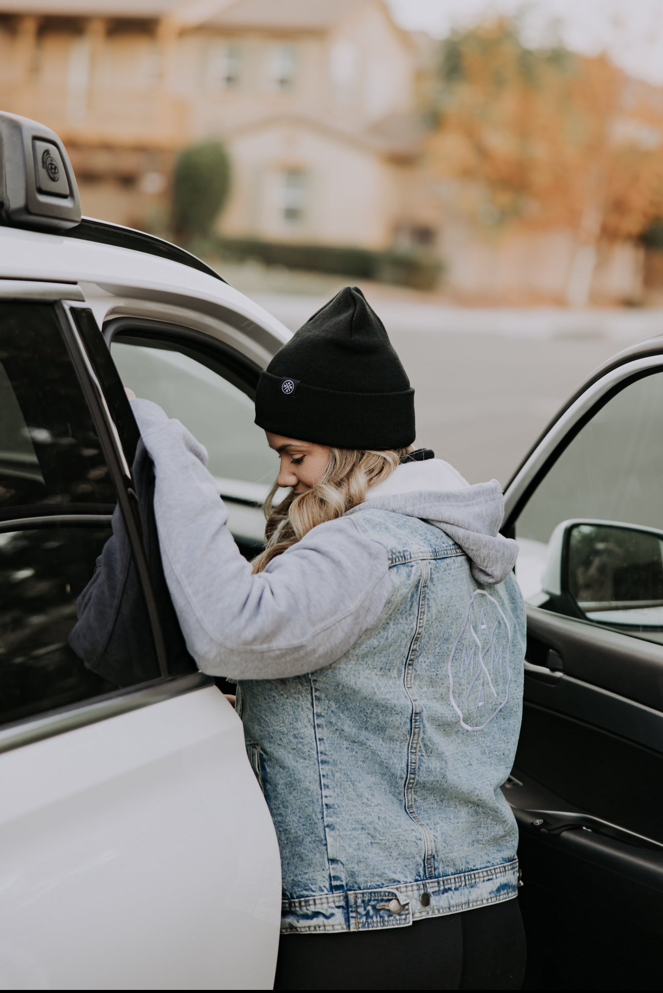 A person is wearing a black beanie and the Mama X™ Brand's Mama X™ Embroidered Denim Jacket with a classic boyfriend fit. They are standing beside an open car door, placing items inside, while the backdrop features a blurred view of trees and houses.