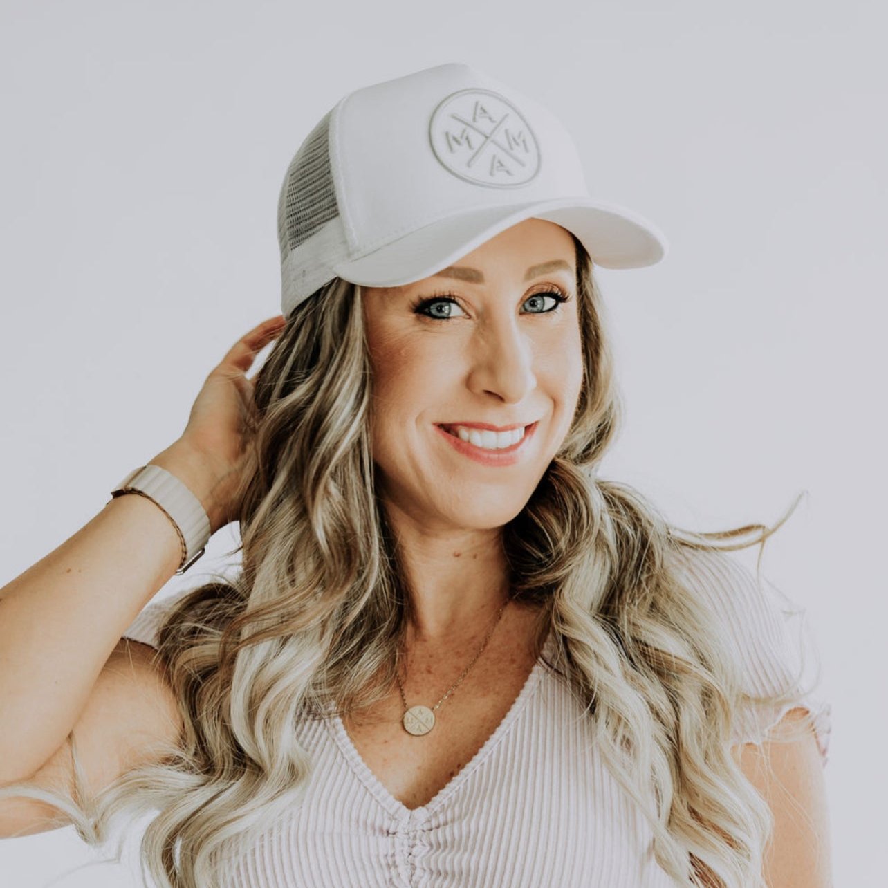 A woman with long, wavy blonde hair smiles while wearing a Mama X™ Premium Canvas Trucker Hat in White/Gray from the Mama X™ Brand, along with a light-colored, short-sleeved shirt. Her left hand gently touches the side of her hat, which features the distinctive MAMA X™ logo. She is adorned with a necklace and bracelets. The background is plain and light-colored.