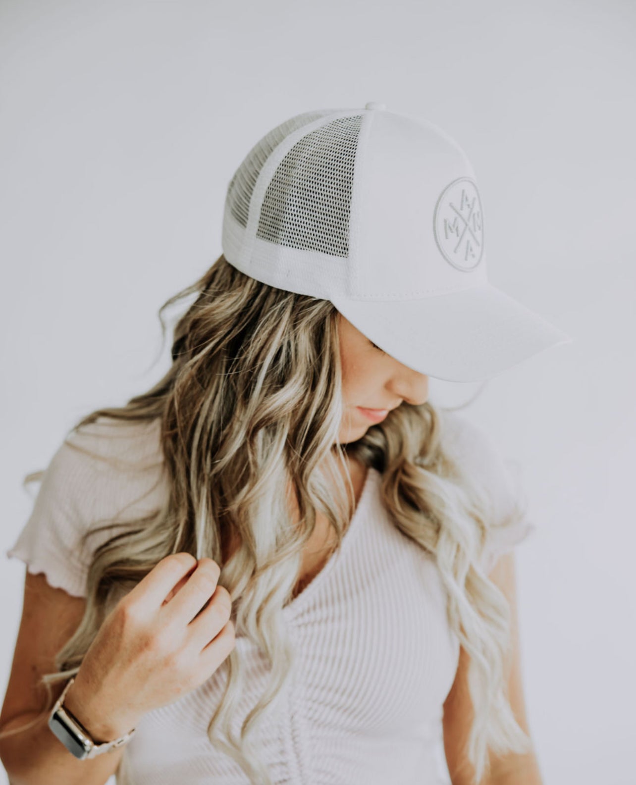 A woman with long, wavy, blonde hair is wearing a Mama X™ Premium Canvas Trucker Hat in White/Gray adorned with the MAMA X™ logo on the front. She is looking downward and has her right hand adjusting the brim of the hat. She wears a light-colored, short-sleeve, ribbed top.