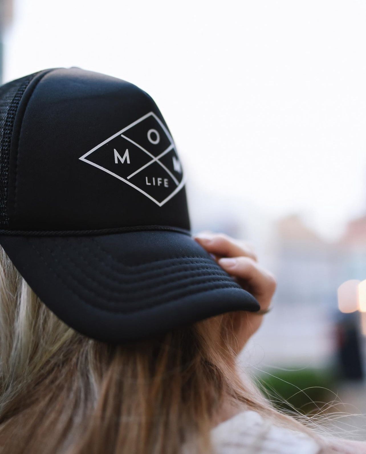 A person with long, light-colored hair is wearing a black Mom Life Foam Hat from Mama X™ Brand, featuring a white diamond-shaped logo. The individual is facing away from the camera, adjusting the hat's plastic adjustable snap. The background is slightly blurred.