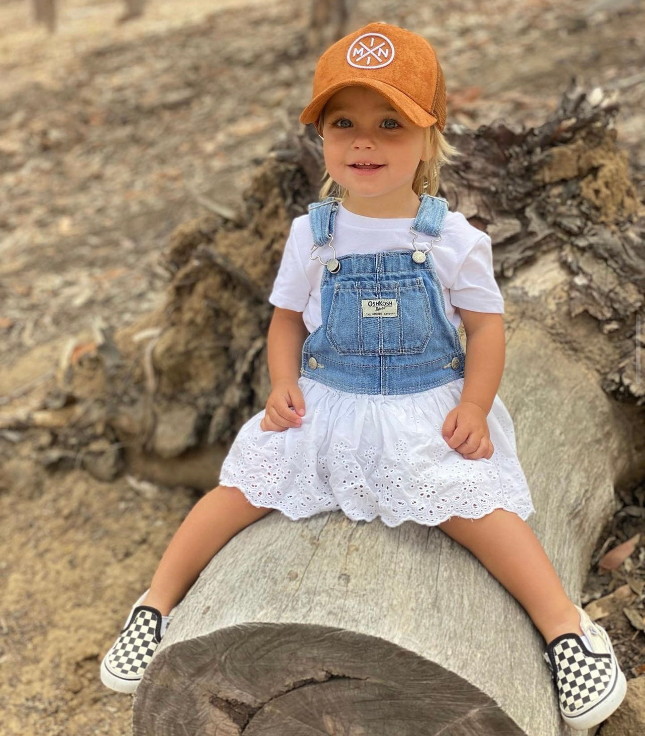 A young child sits on a large log outdoors, wearing the cutest hat—a Mini X Brown Suede Trucker Hat from Mama X™ Brand—paired with a white t-shirt, denim overalls with a white lace skirt underneath, and black-and-white checkered slip-on shoes. The background reveals a natural setting with dried foliage and wood, perfect for a fall outfit.