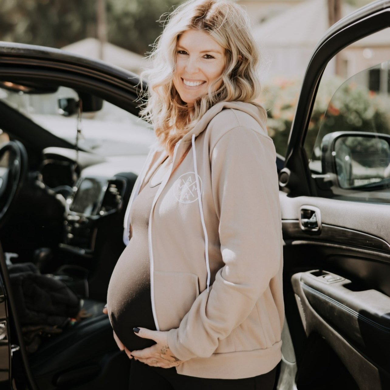 A pregnant woman stands next to an open car door, wearing a brown Mama X™ Brand zip-up sweatshirt and black shorts, smiling at the camera. She has long, wavy blonde hair and is holding her belly with both hands. The background shows trees and a building.