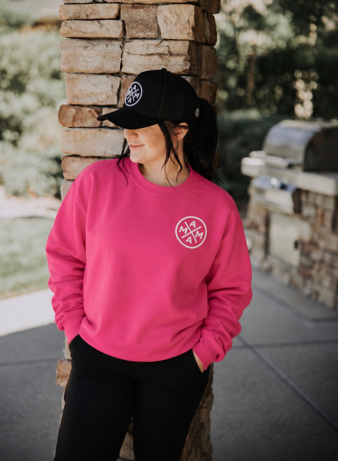 A person wearing a bright pink Hot Pink Mama X Crewneck with a circular white logo on the chest and a matching black baseball cap.