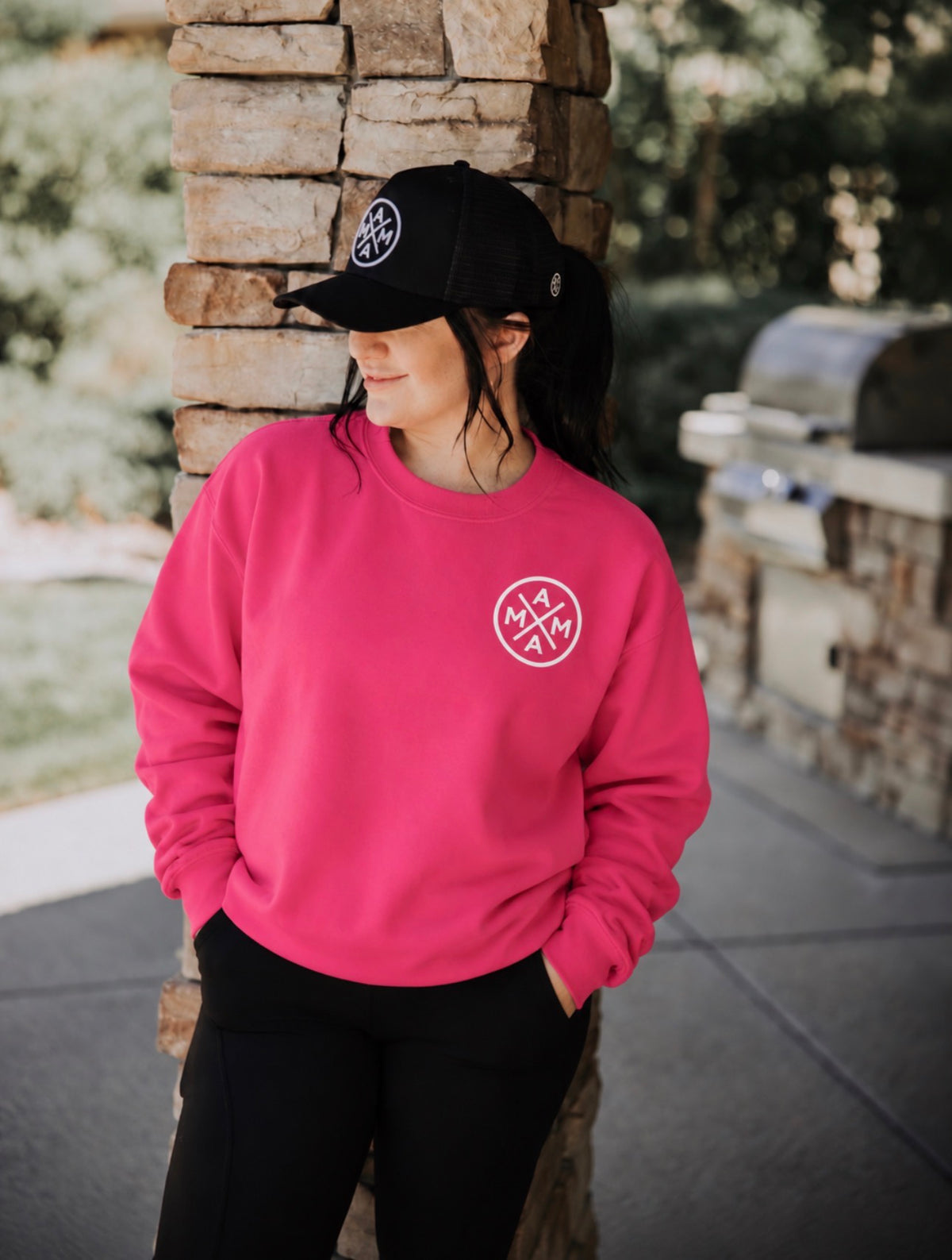 A woman wearing a hot pink Mama X Crewneck and a black baseball cap, both featuring a circular white logo.