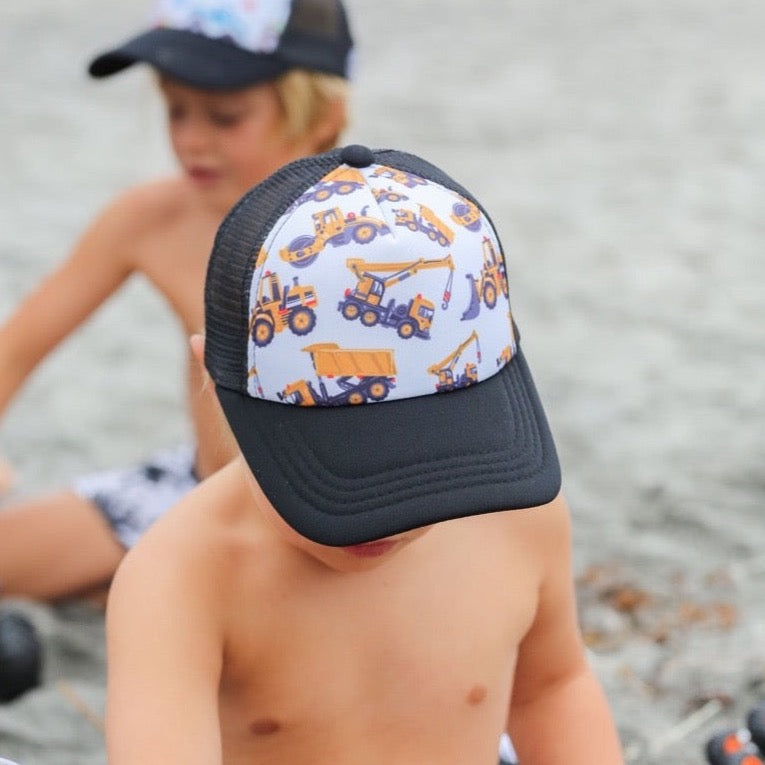 Boy playing in the sand on the beach wearing a black trucker hat with white canvas and yellow tractors 