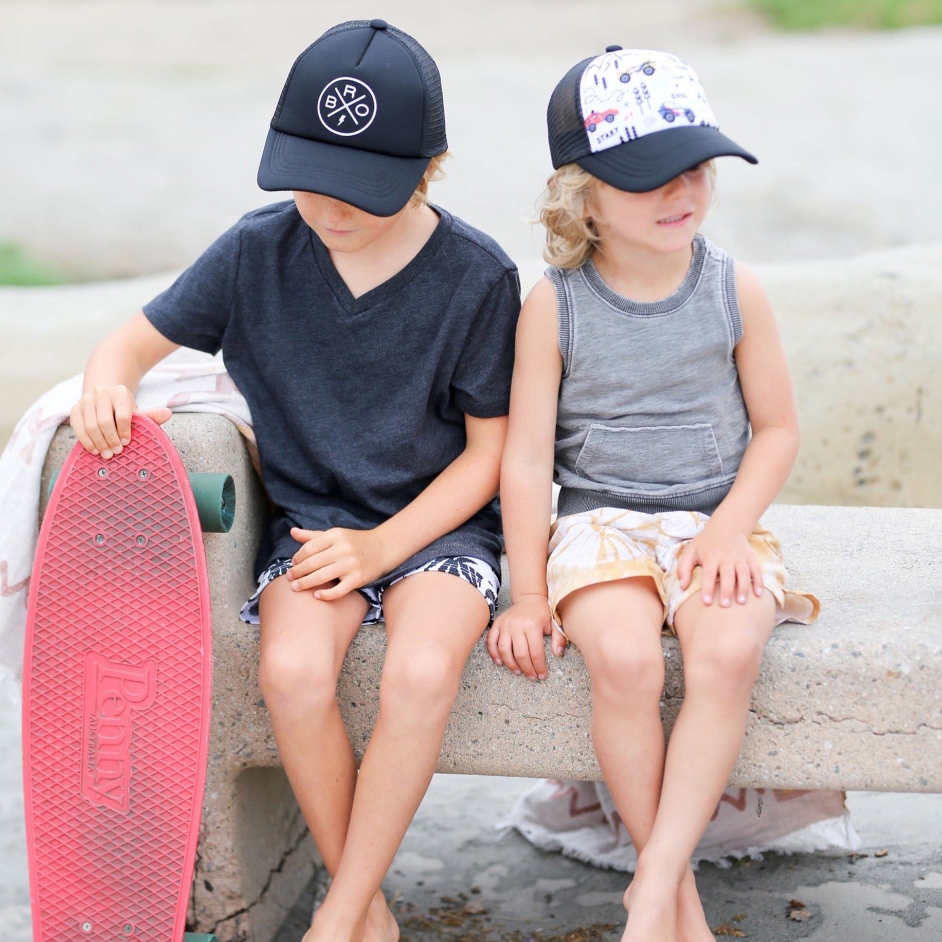 Two young children are sitting on a stone bench, both stylishly sporting Mama X™ Brand's Bro X Trucker Hats and casual tops in Baby Toddler Youth sizes. One child is holding a red skateboard, while the other has their hands resting on their lap. The background showcases a blurred outdoor setting, ensuring they are well-protected from the sun with their Bro X Trucker Hats.