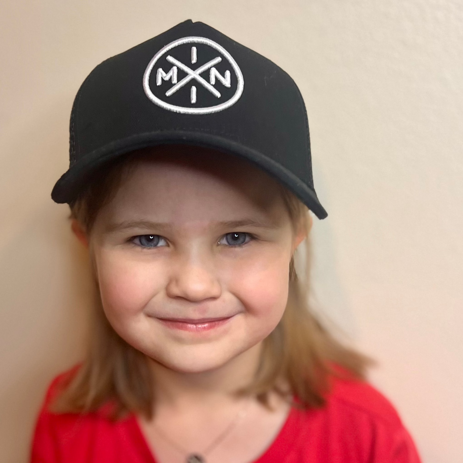 A young child with light brown hair smiles while wearing the Mini X Premium Trucker Hat from Mama X™ Brand. The black cap is adorned with a circular white embroidered logo featuring the letters "M," "I," and "N." The child, dressed in a red shirt, stands against a plain beige background.