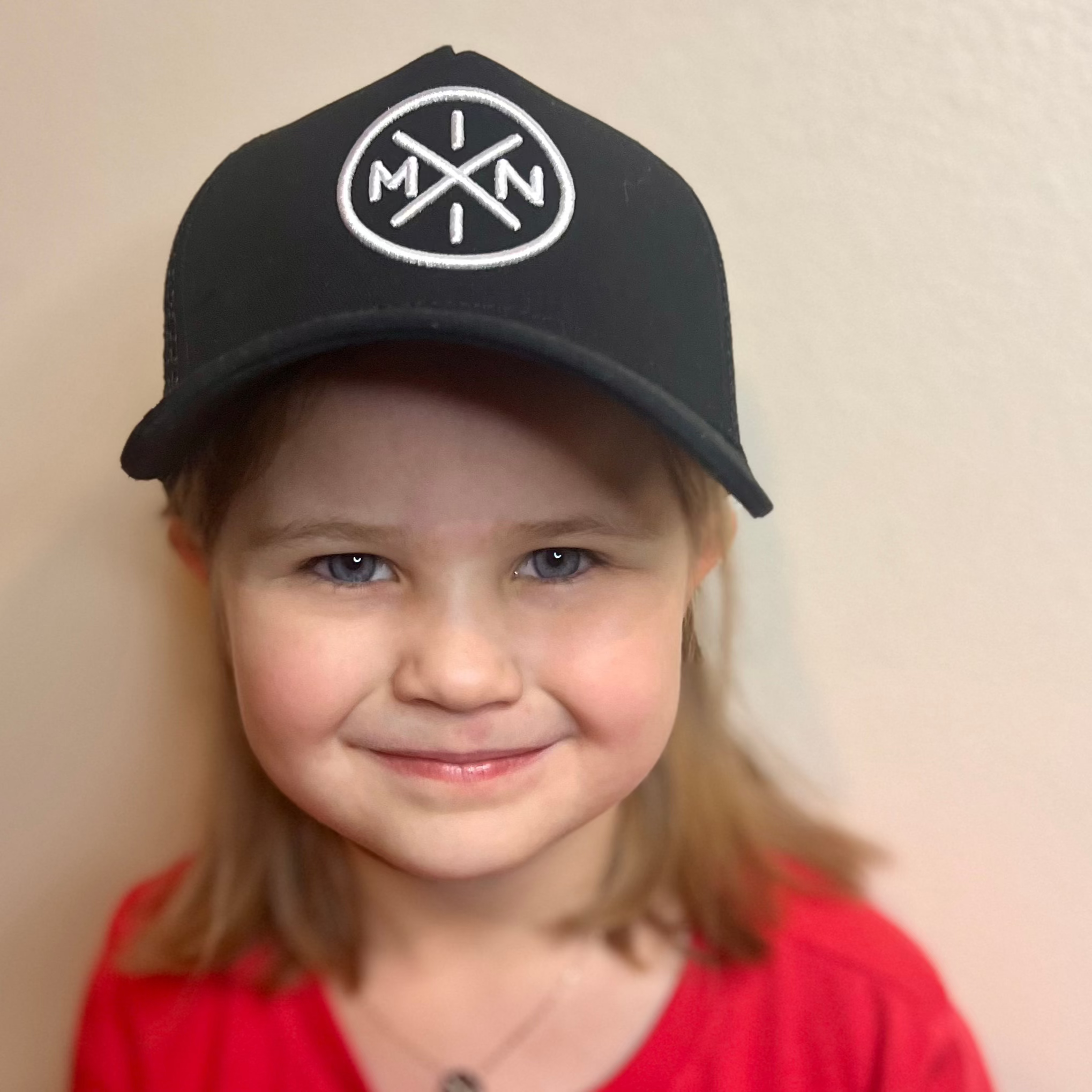 A young child with light brown hair smiles while wearing the Mini X Premium Trucker Hat from Mama X™ Brand. The black cap is adorned with a circular white embroidered logo featuring the letters 