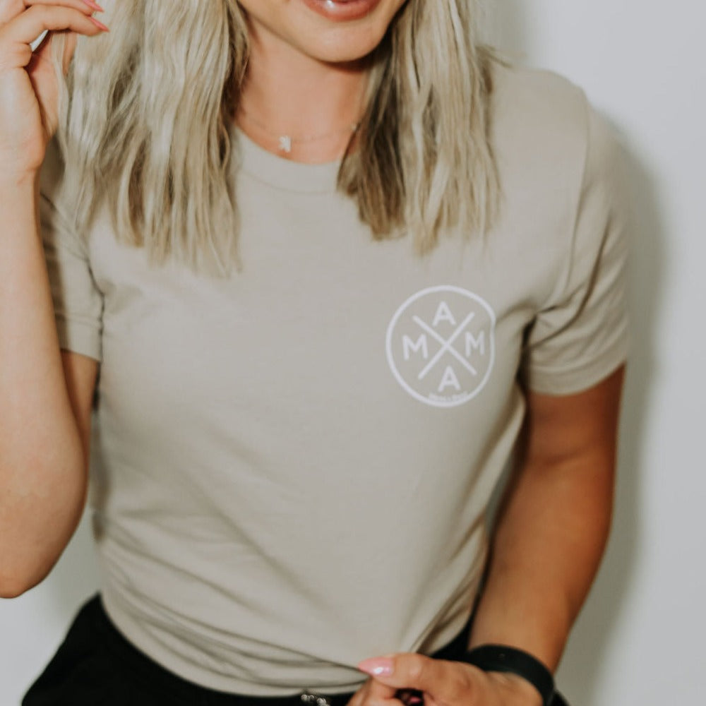 A person with blonde hair is wearing a Mama X™ T-Shirt - Sand featuring the circular 'Mama X™' logo on the front. Their hand is partially raised, holding a strand of hair, against a plain light-colored background.