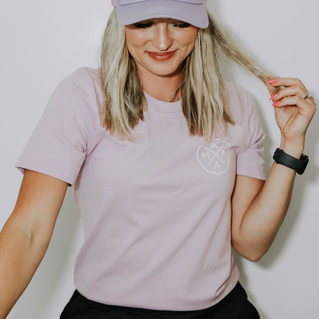 A woman with long blonde hair wearing a light lilac Mama X™ T-Shirt from Mama X™ Brand in unisex sizing and a matching cap featuring a small logo on the front. She is smiling slightly, touching a strand of her hair with one hand. She has bright pink nails and is also wearing a black watch.