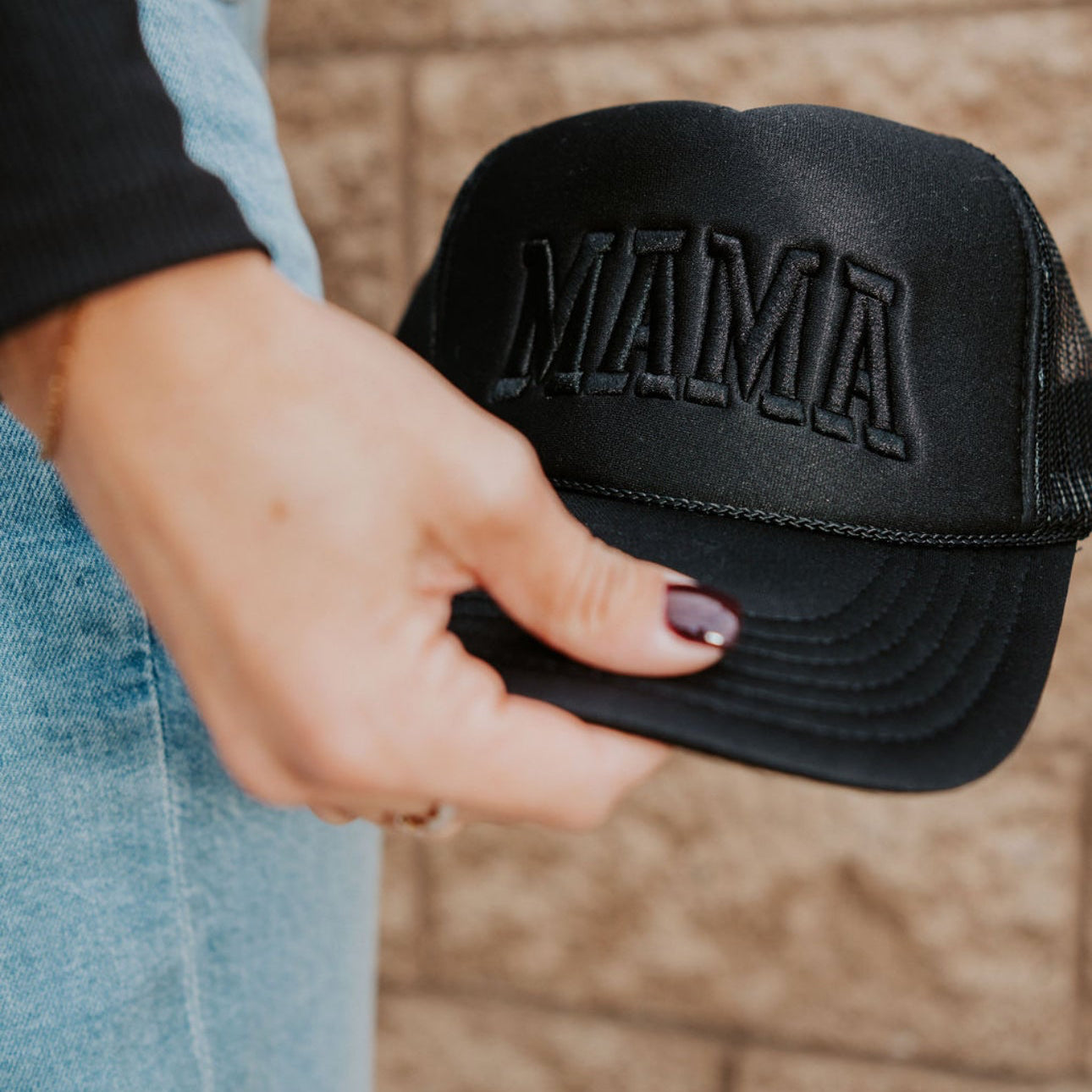 A person with painted nails holds a Black out foam Trucker cap by Mama X™ Brand, featuring "MAMA" embroidered on the front, standing before a textured beige wall.
