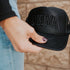 A person with painted nails holds a Black out foam Trucker cap by Mama X™ Brand, featuring "MAMA" embroidered on the front, standing before a textured beige wall.