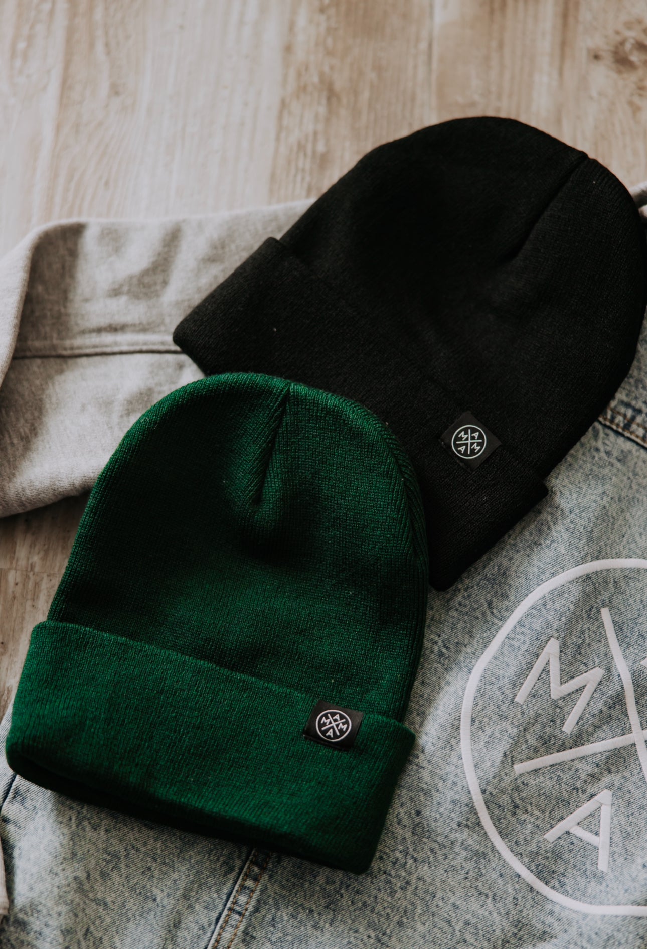 Two beanies from the Mama X™ Brand are displayed on clothing. The black Mama X™ Cuffed Beanie with a 12-inch cuffed design rests on a light gray garment, while the green Mama X™ Cuffed Beanie sits on a denim jacket, featuring a white circular logo. Both beanies showcase a small round emblem.