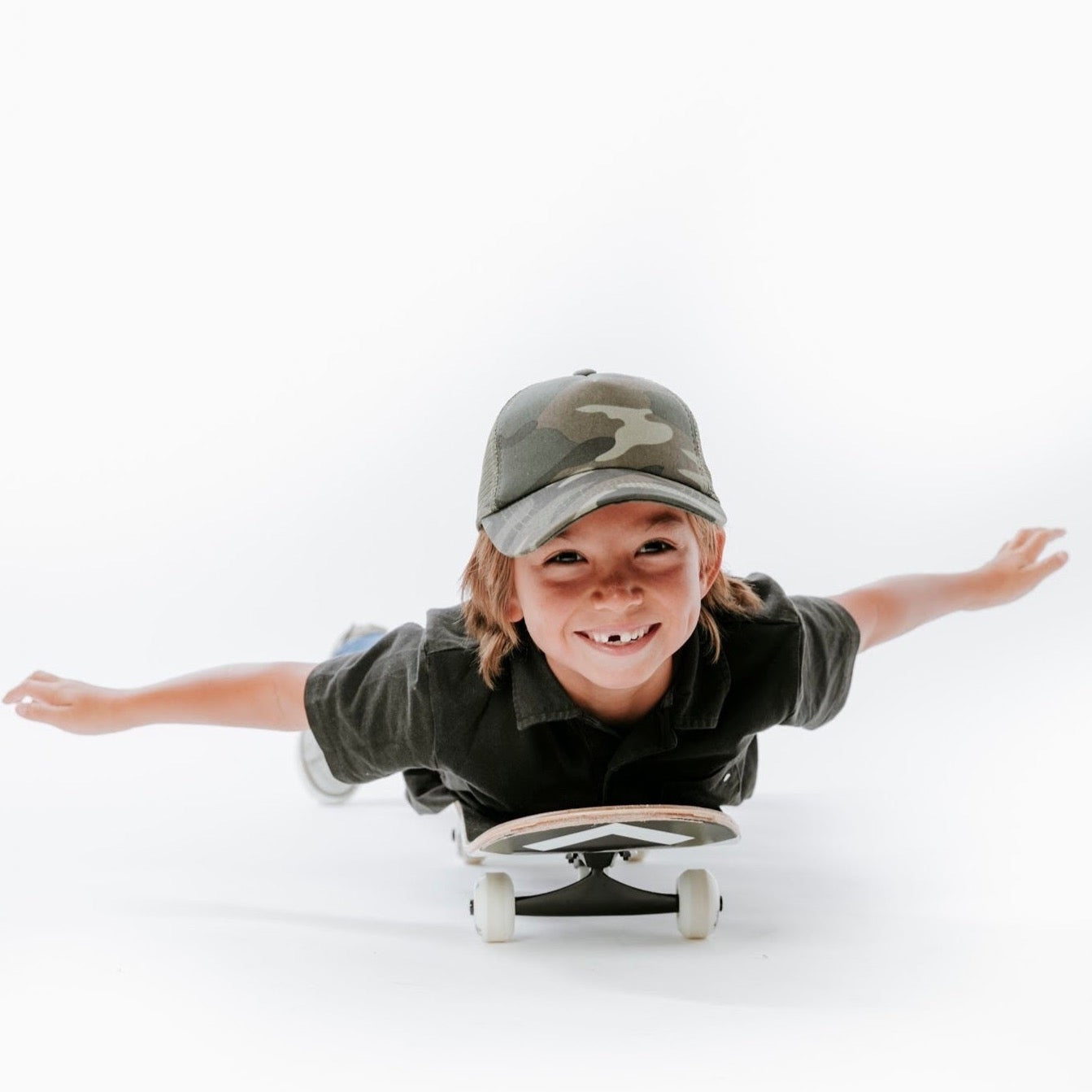 A cheerful young boy, sporting a Mama X™ Brand Camo Trucker Hat and a black shirt, lies face down on a skateboard with his arms spread wide as if flying. The plain white background accentuates his joyful expression and playful pose, making them the main focus of the image.