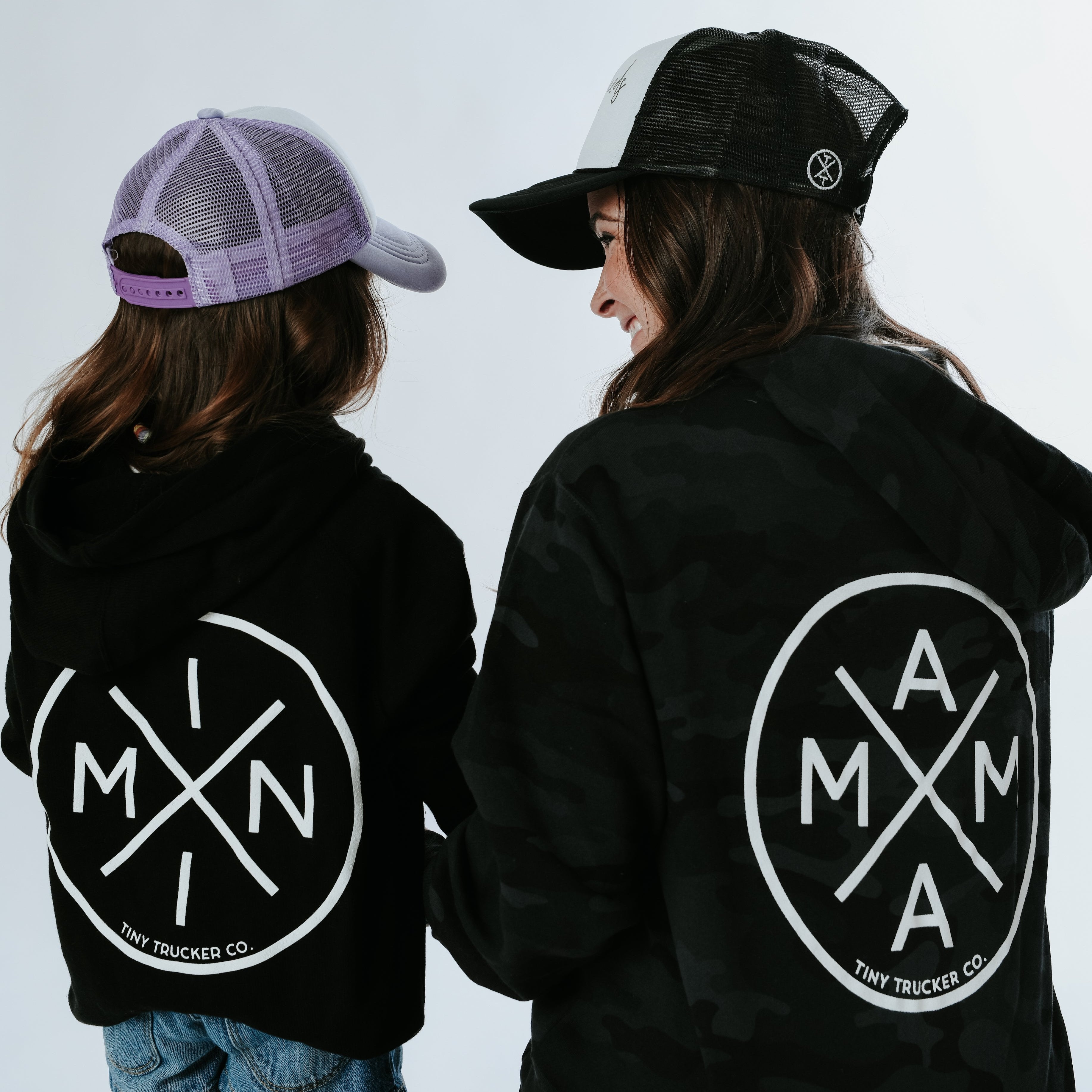 A woman and child are facing away from the camera, both wearing dark Mama X™ Brand hoodies with zip ups and trucker hats. Their hoodies feature the same circular Mama X™ logo with the letters 