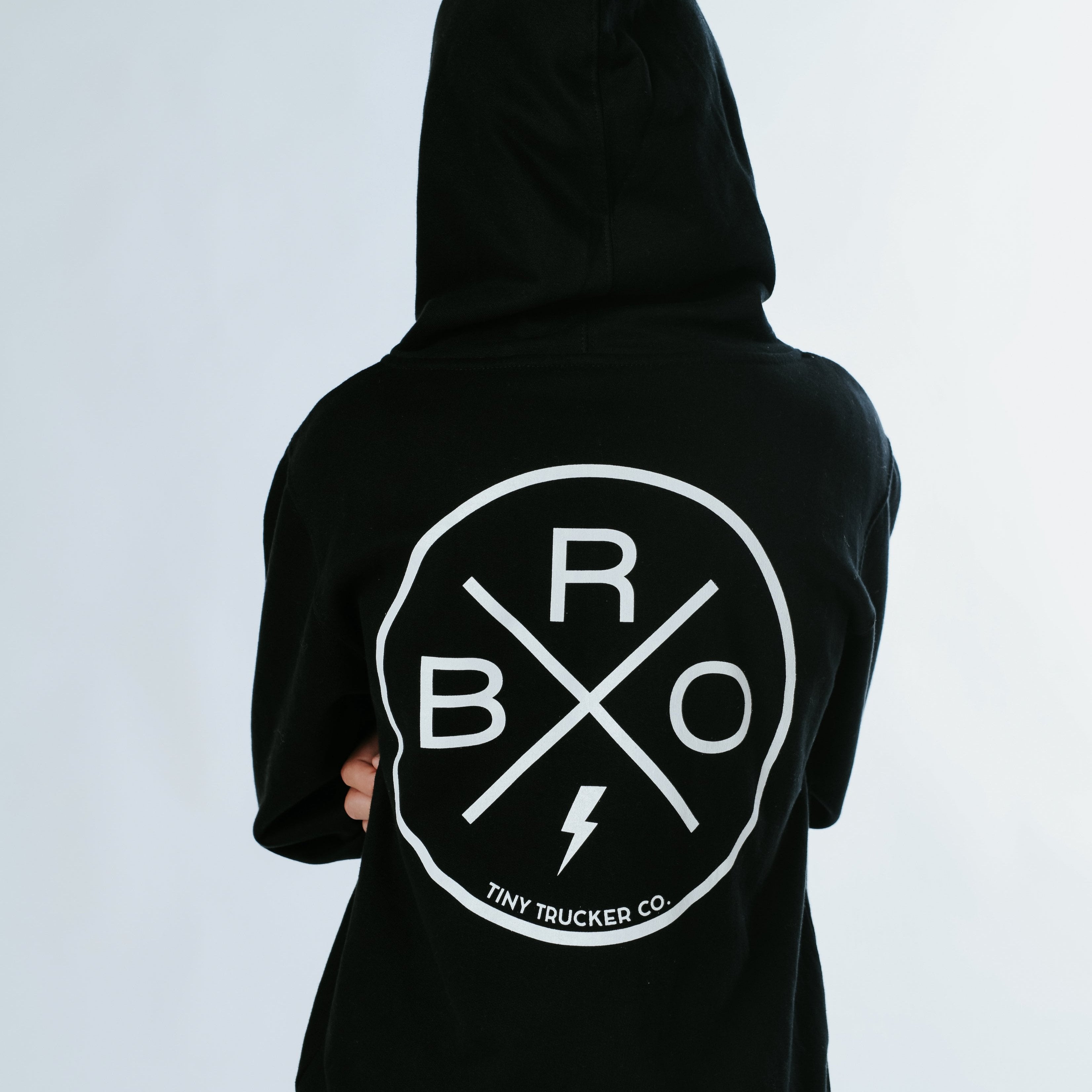 A person is facing away from the camera wearing the Bro X Zip Up - Tiny Trucker hoodie by Mama X™ Brand, with the hood up. The back of the hoodie features a white circular logo with the letters 