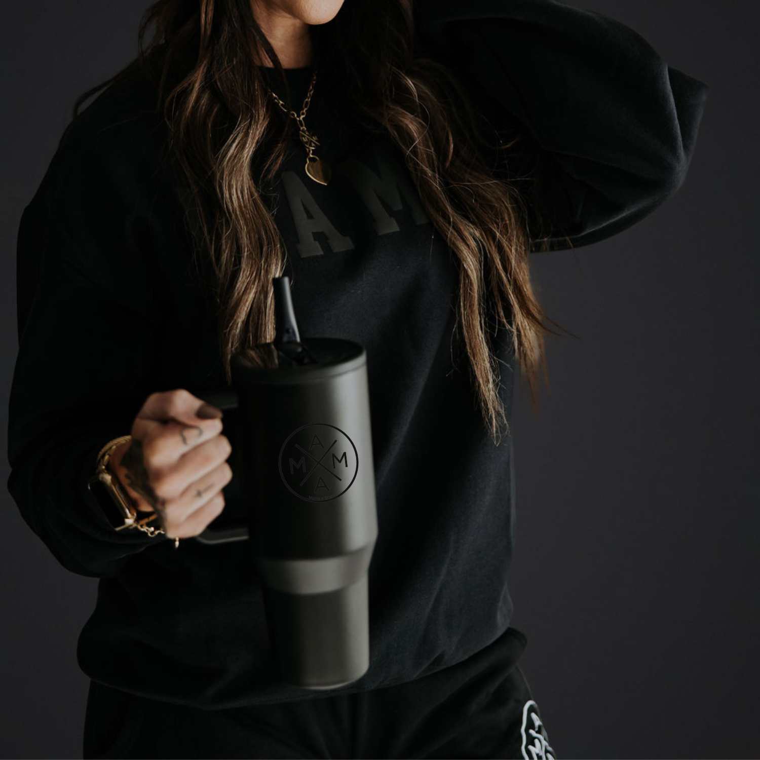 A person wearing a black sweatshirt holds a **Blacked-Out Mama X™ Tumbler** featuring the **Mama X™ Brand** logo and a straw. The background is dark, highlighting their necklace with a pendant, which complements their casual, modern style.