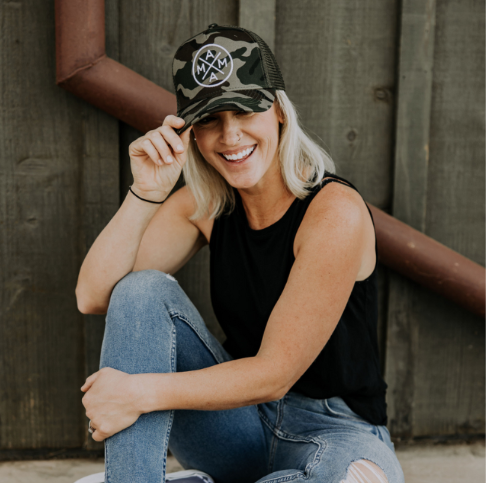 A person with shoulder-length blonde hair sits against a wooden wall, smiling. They wear a Mama X™ Brand Premium Canvas Trucker Hat in camo, a sleeveless black top, and ripped jeans, holding the brim of the hat with one hand. A rust-colored pipe is visible behind them.