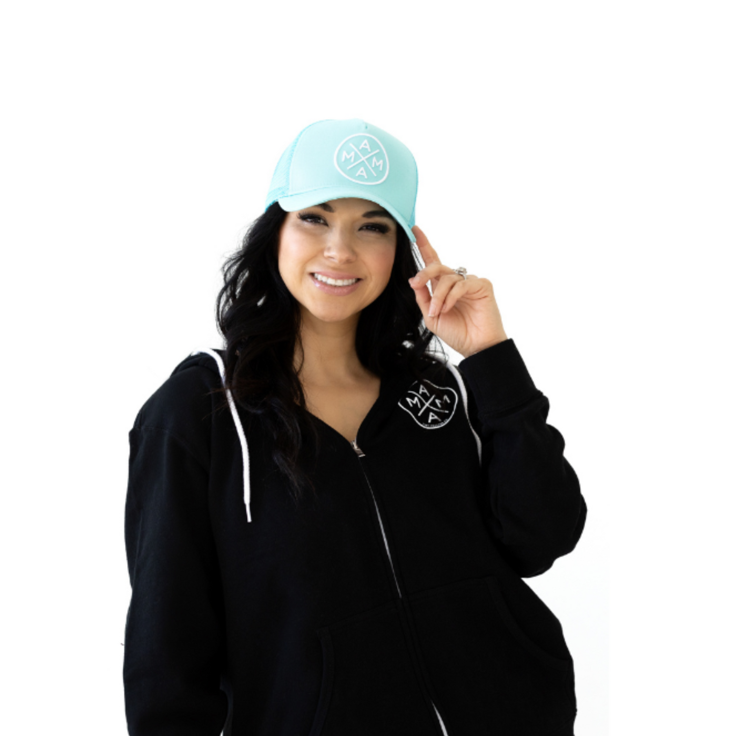 A smiling woman with long dark hair wears the Mama X™ Premium Canvas Trucker Hat in mint and a black zip-up hoodie. She holds the brim of the 5-panel cap with her right hand. The cap and hoodie both feature a circular MAMA X logo. The background is white.