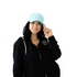 A smiling woman with long dark hair wears the Mama X™ Premium Canvas Trucker Hat in mint and a black zip-up hoodie. She holds the brim of the 5-panel cap with her right hand. The cap and hoodie both feature a circular MAMA X logo. The background is white.