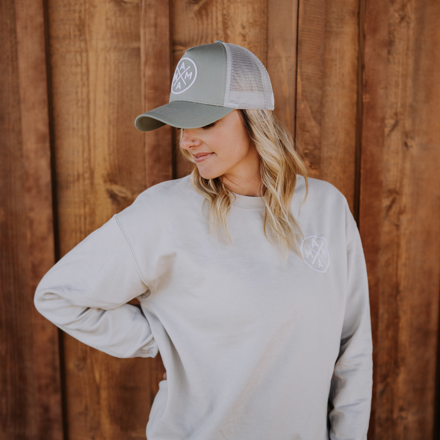 A person with long blonde hair, wearing a light gray sweatshirt and the Mama X™ Premium Canvas Trucker Hat in Sage from the Mama X™ Brand, stands against a wooden background with their head turned slightly to the side while looking down.