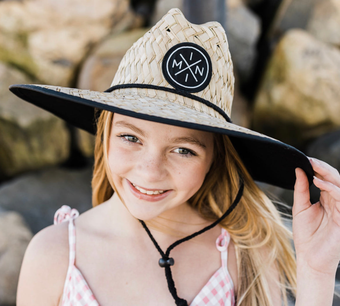 A young girl with long blonde hair smiles while wearing the Mini X Lifeguard Hat - Toddler/Youth from Mama X™ Brand—a wide-brimmed straw hat with a black band and round logo. She is outdoors, against a stone background, dressed in a pink and white checkered top with thin straps.