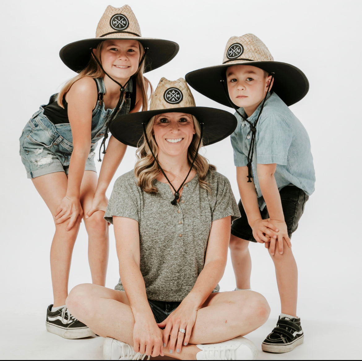 A woman sits cross-legged on the floor, smiling, with a young girl and boy standing on either side. All are wearing Mama X™ Brand Mini X Lifeguard Hats - Toddler/Youth for sun protection. The girl is dressed in denim overalls, the boy in a blue shirt and shorts, and the woman in a gray shirt and shorts.