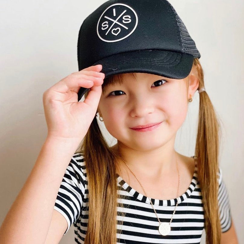 A young girl wearing a Mama X™ Brand Sis X Trucker Hat with a white logo and a black and white striped shirt smiles and holds the brim of her 5-panel cap. She has two long pigtails and is accessorized with gold earrings and a necklace with a circular pendant.