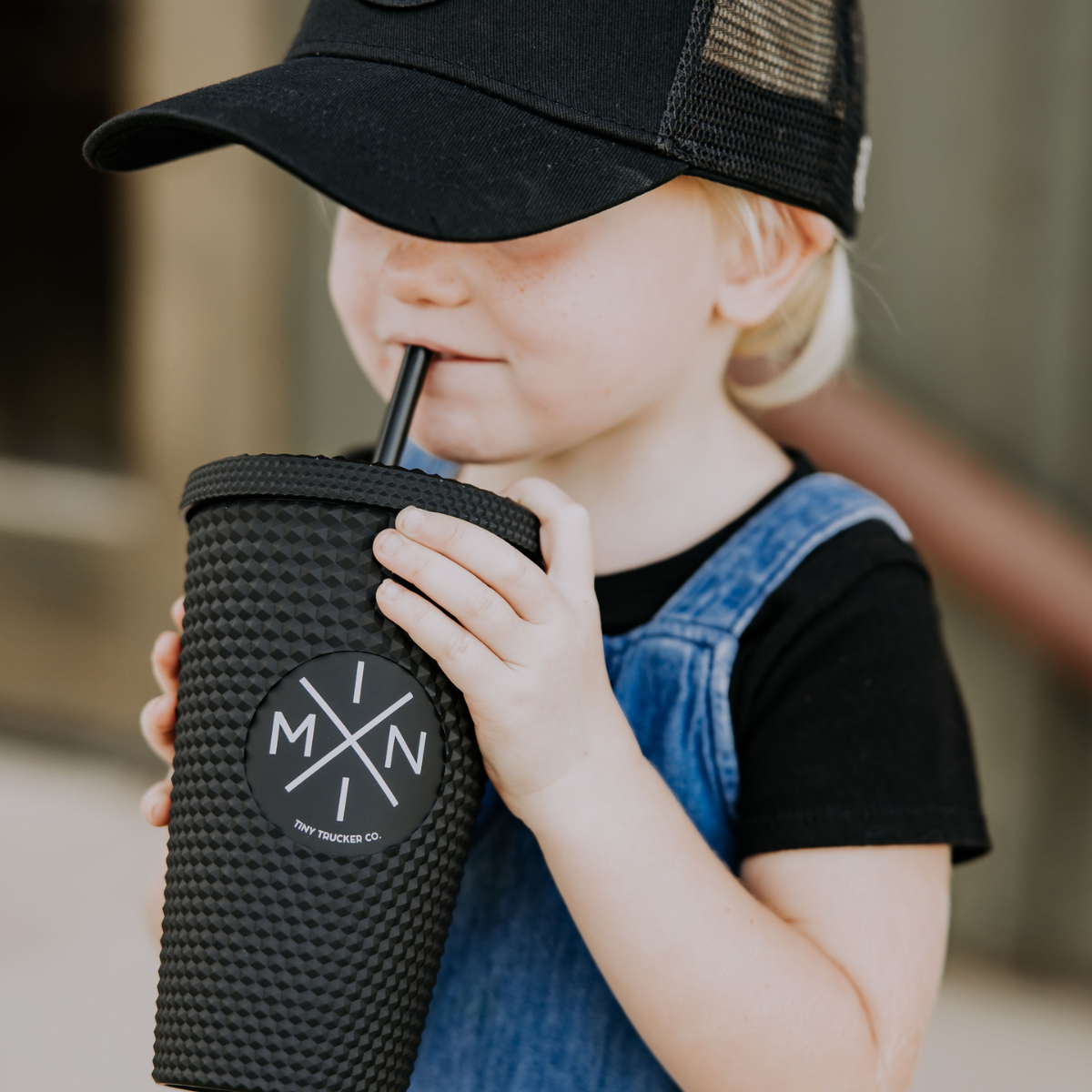 A young child in a black cap and denim overalls drinks from a 16oz Mama X™ Brand Mini X Studded Plastic Tumbler with a reusable straw. The tumbler features a round label with the letters "M," "I," "N," and "I" arranged in a cross pattern on the front. The background is blurred.