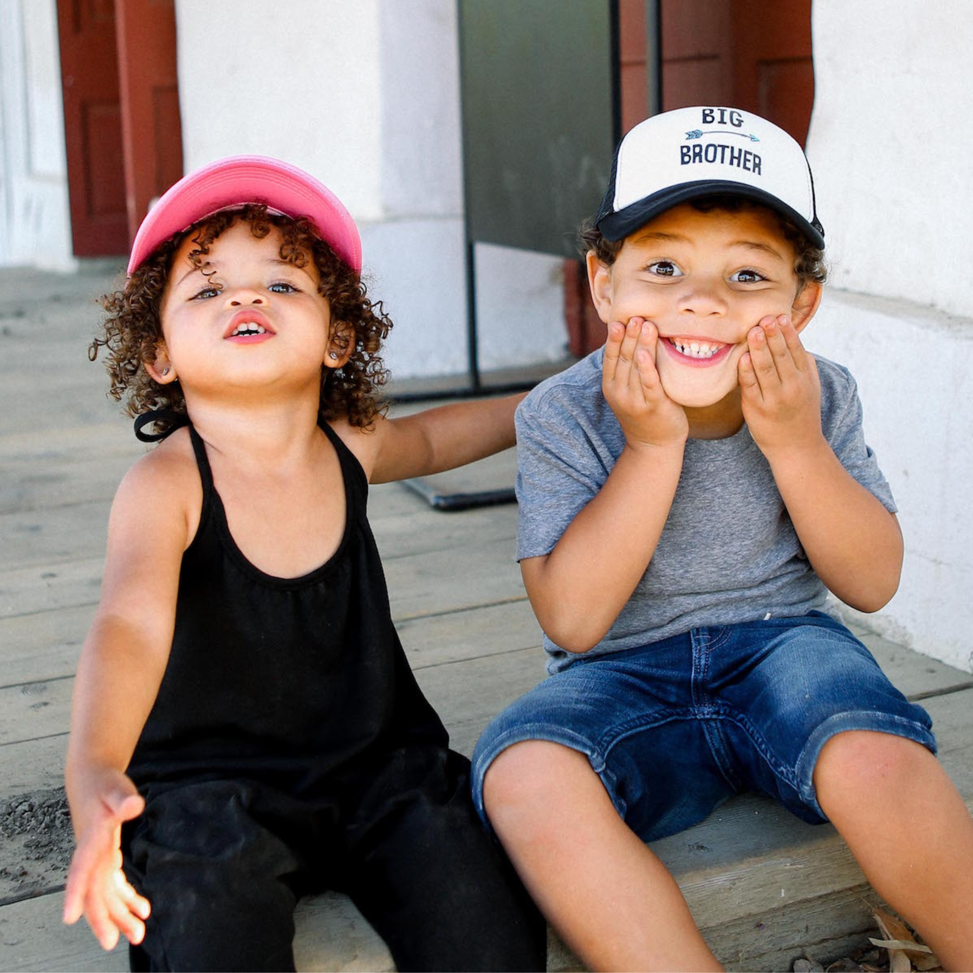 Two young children are sitting on a wooden porch. The child on the left, wearing a pink baseball cap and black tank top, has curly hair and an open-mouthed expression. The child on the right, sporting a 