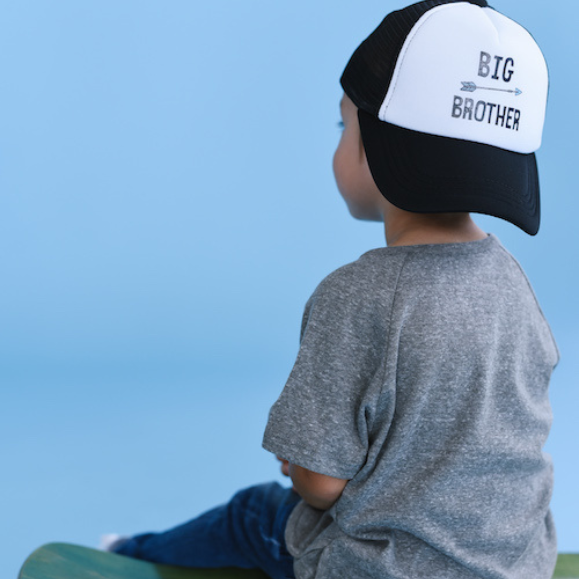 A young child wearing a grey t-shirt and a high-quality Mama X™ Brand Big Brother Trucker Hat sits on a green stool, facing away from the camera against a light blue background.