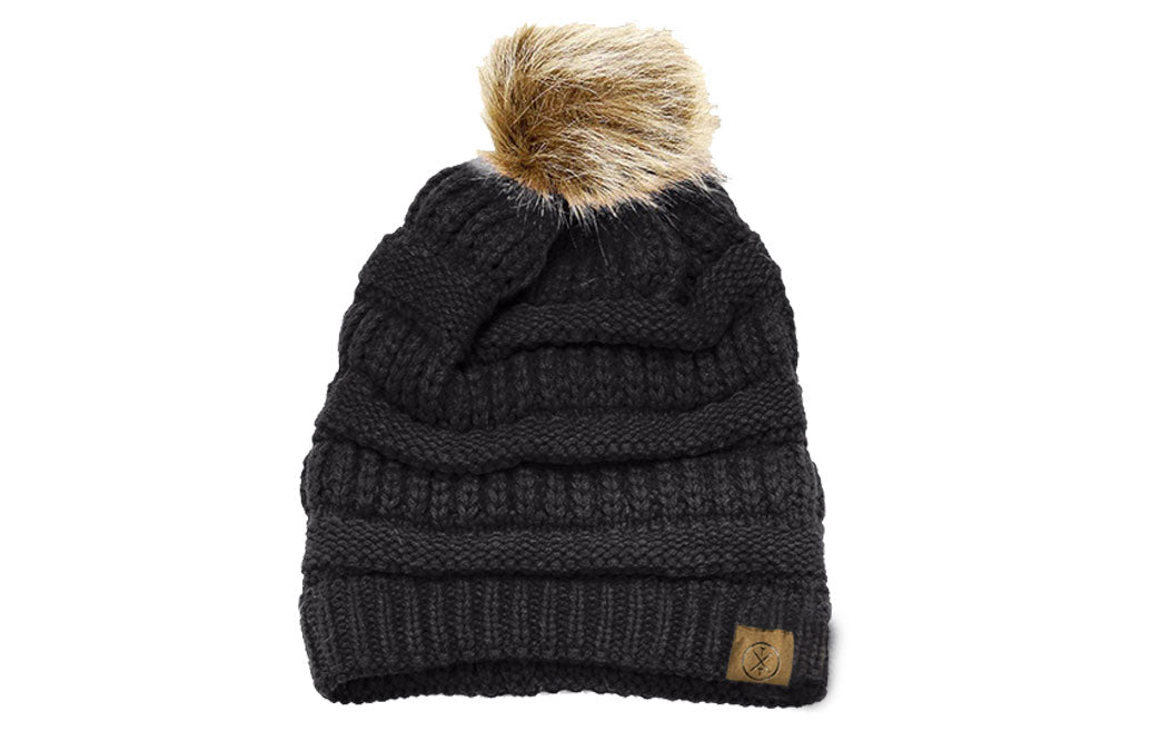 Introducing the **Child Pom Beanie - Black** by Mama X Brand: a stylish black knit beanie featuring a light tan faux fur pom-pom on top. Its ribbed texture and small brown patch on the brim effortlessly blend style with comfort.