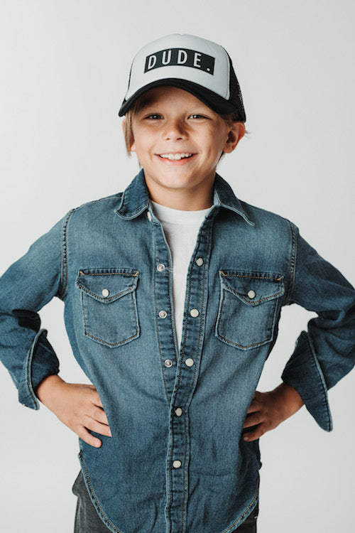 A young boy smiles confidently with his hands on his hips, wearing a denim shirt over a white t-shirt and a Mama X™ Brand Dude Trucker Hat. The background is plain white.