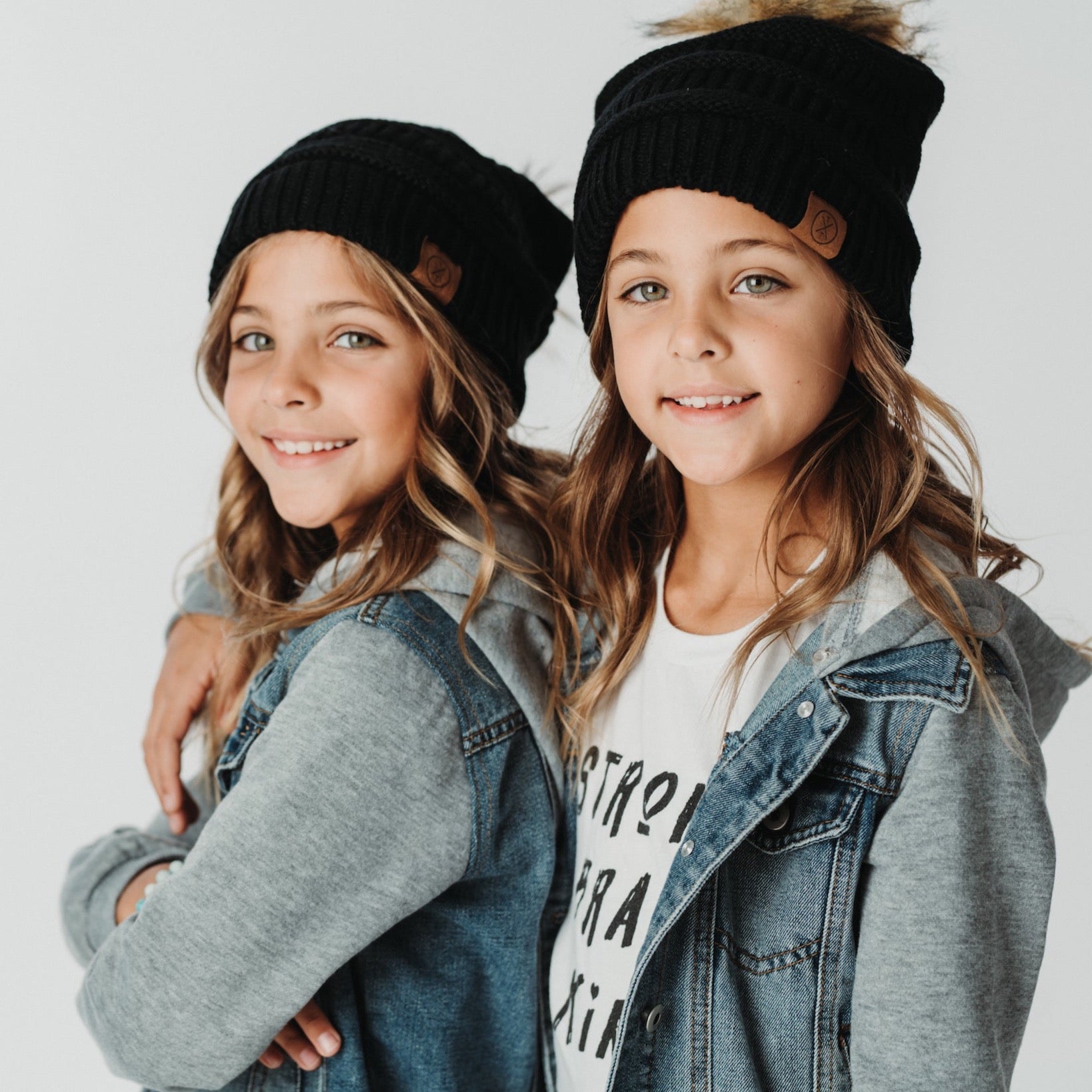 Two young girls stand side by side, each wearing the Child Pom Beanie in black from Mama X Brand, denim jackets, and gray hoodies. They are smiling at the camera against a plain white background.