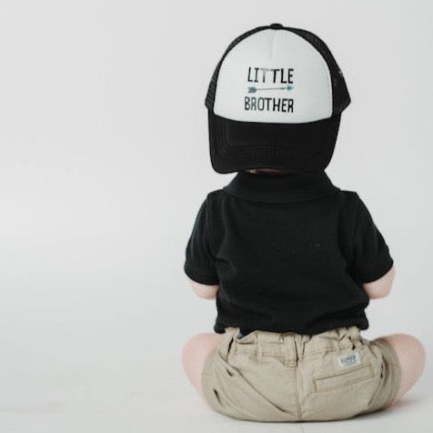 A toddler, seen from behind, is wearing a "Little Brother Trucker Hat" by Mama X™ Brand. The child is dressed in a black shirt and beige shorts, sitting against a plain white background—a perfect moment for family photos or gender reveals.