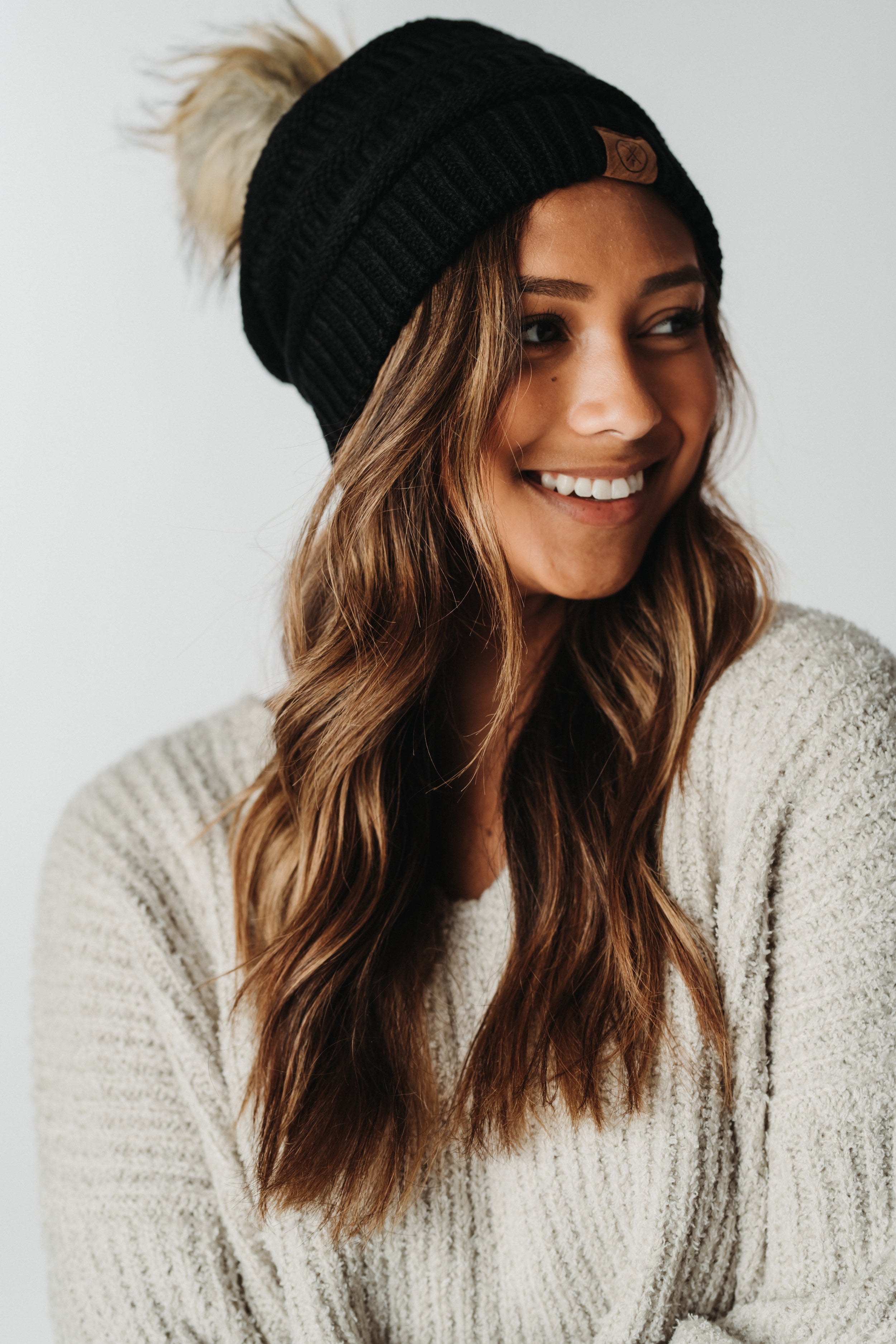 A smiling person with long wavy hair wears a cozy beige sweater and the Mama X Brand Child Pom Beanie in black, featuring a faux fur pom, against a neutral background.