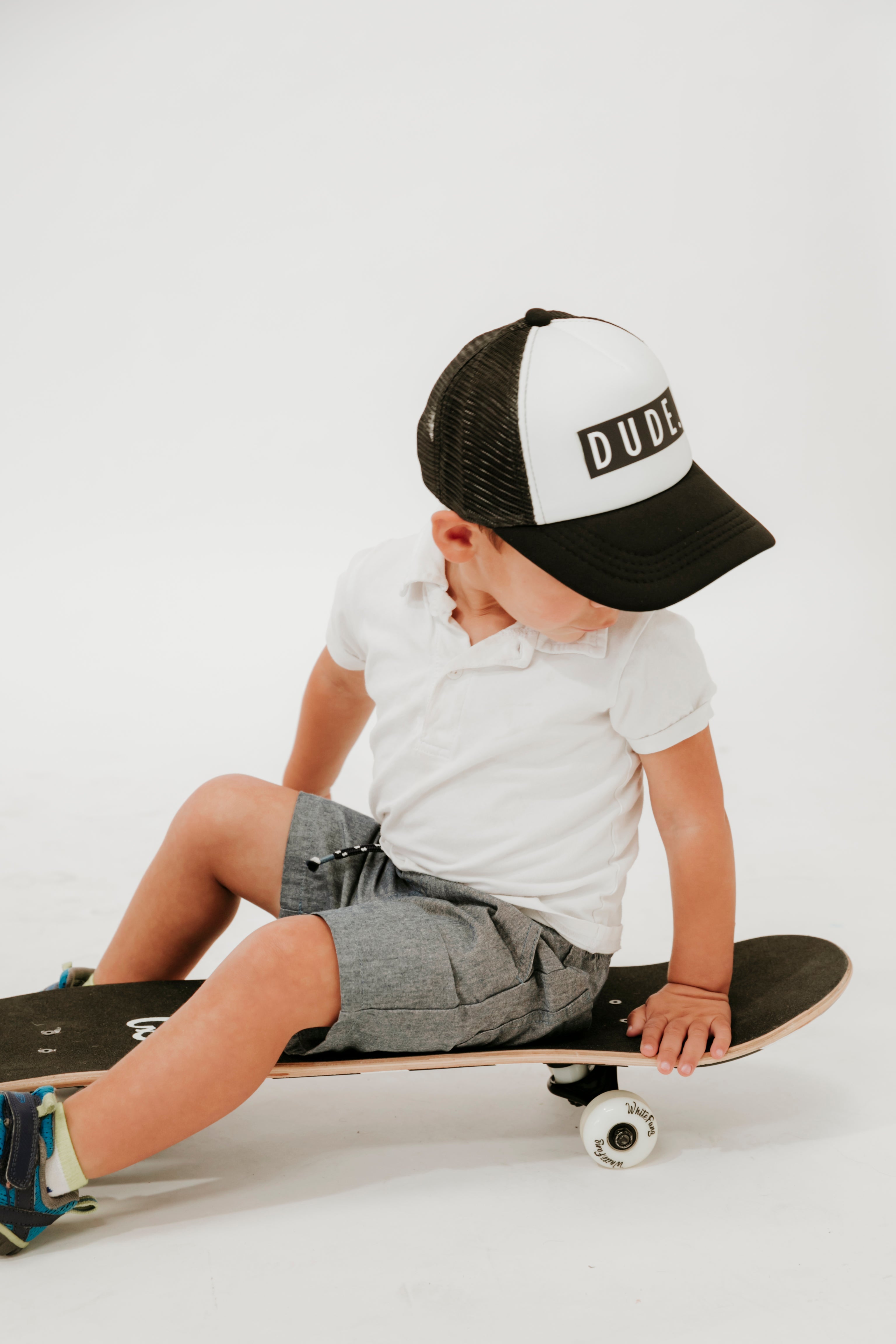 A young child sits on a skateboard in a white studio setting, facing slightly to the right. They are wearing a black and white Mama X™ Brand Dude Trucker Hat with the word "DUDE" on it, a white polo shirt, gray shorts, and black sneakers. Their hand rests on the skateboard for balance.