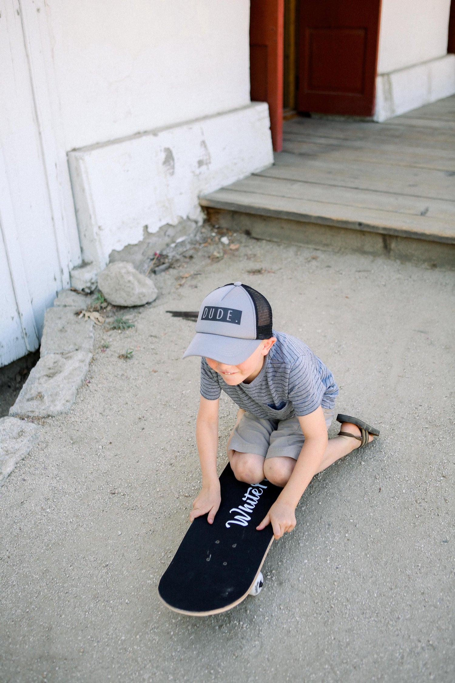 A young boy wearing a "DUDE Trucker Hat - Grey &amp; Black" from Mama X™ Brand and a striped shirt sits on a skateboard, resting on the ground near a wooden porch with steps. He appears to be preparing to ride or taking a break. The environment is outdoors with dirt and some rocks around.