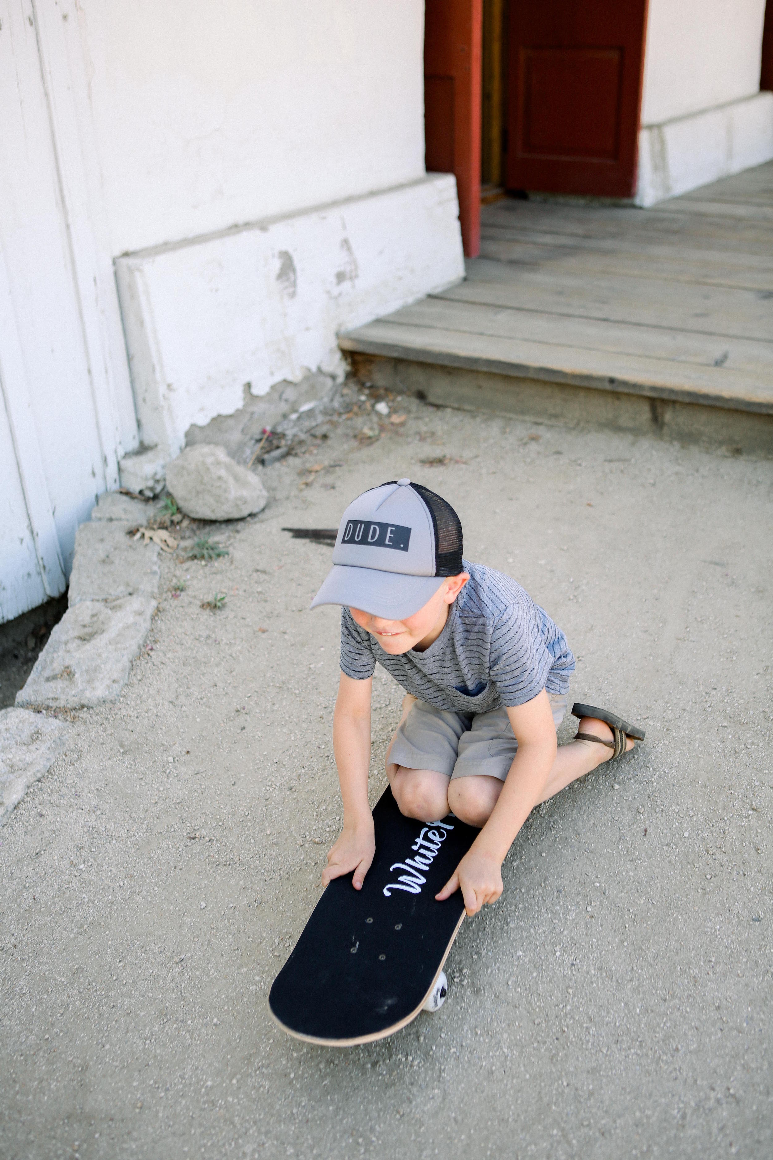 A young boy wearing a "DUDE Trucker Hat - Grey &amp; Black" from Mama X™ Brand and a striped shirt sits on a skateboard, resting on the ground near a wooden porch with steps. He appears to be preparing to ride or taking a break. The environment is outdoors with dirt and some rocks around.