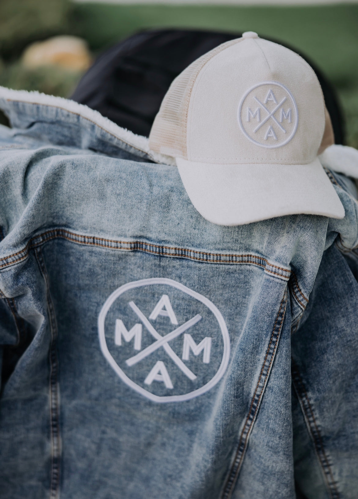 A denim jacket with "MAMA" printed in a circular design on the back is draped over a chair. The Mama X™ Light Suede Trucker Hat from Mama X™ Brand, featuring the same "MAMA" circular design logo, is placed on top of the jacket. The background is blurred greenery.