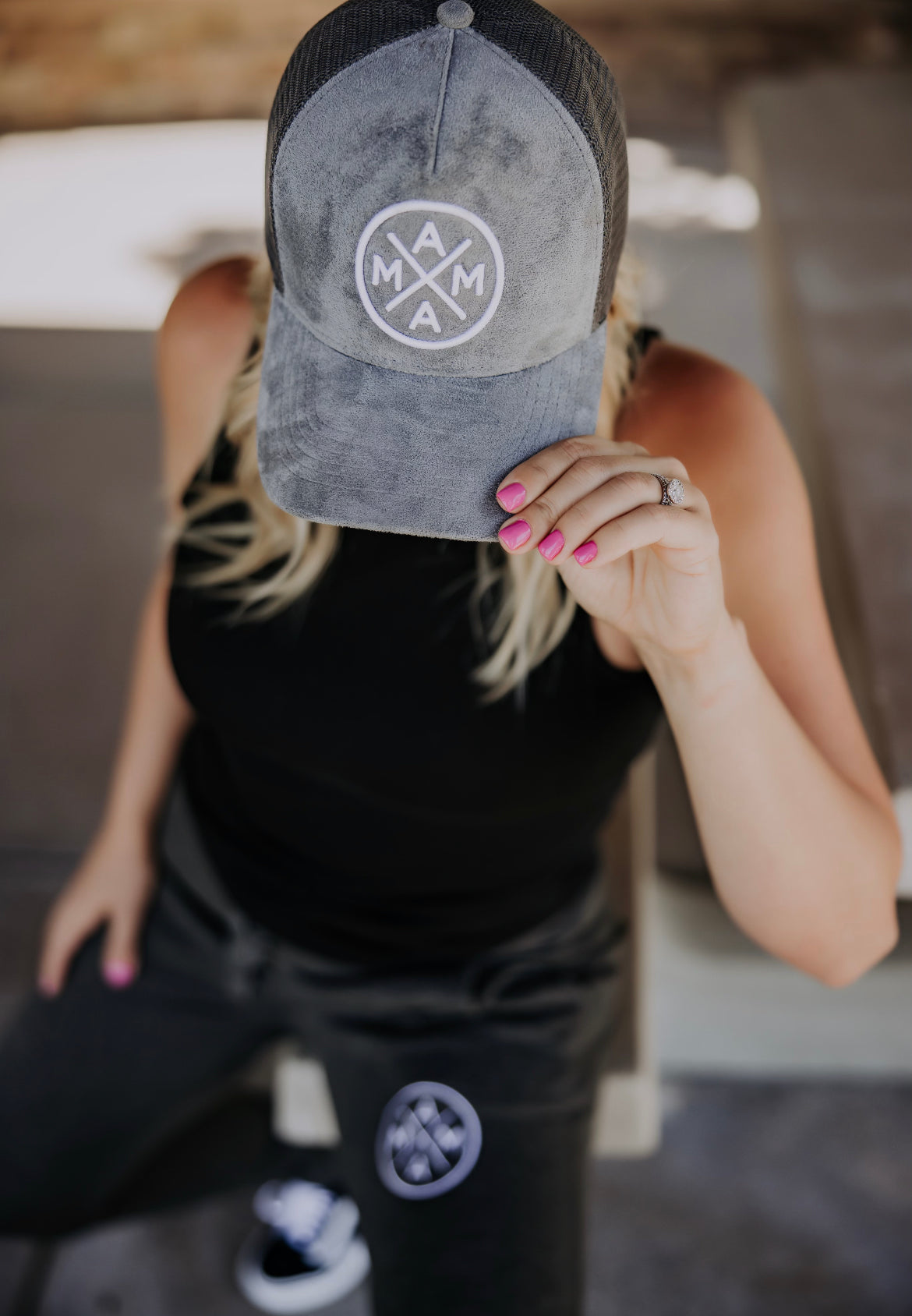 A person with blonde hair is seated, wearing a black sleeveless top and grey pants. They are holding a Mama X™ Trucker Hat - Gray Suede from the Mama X™ Brand over their face, obscuring their identity. Their nails are painted pink, and they have a ring on their left hand.
