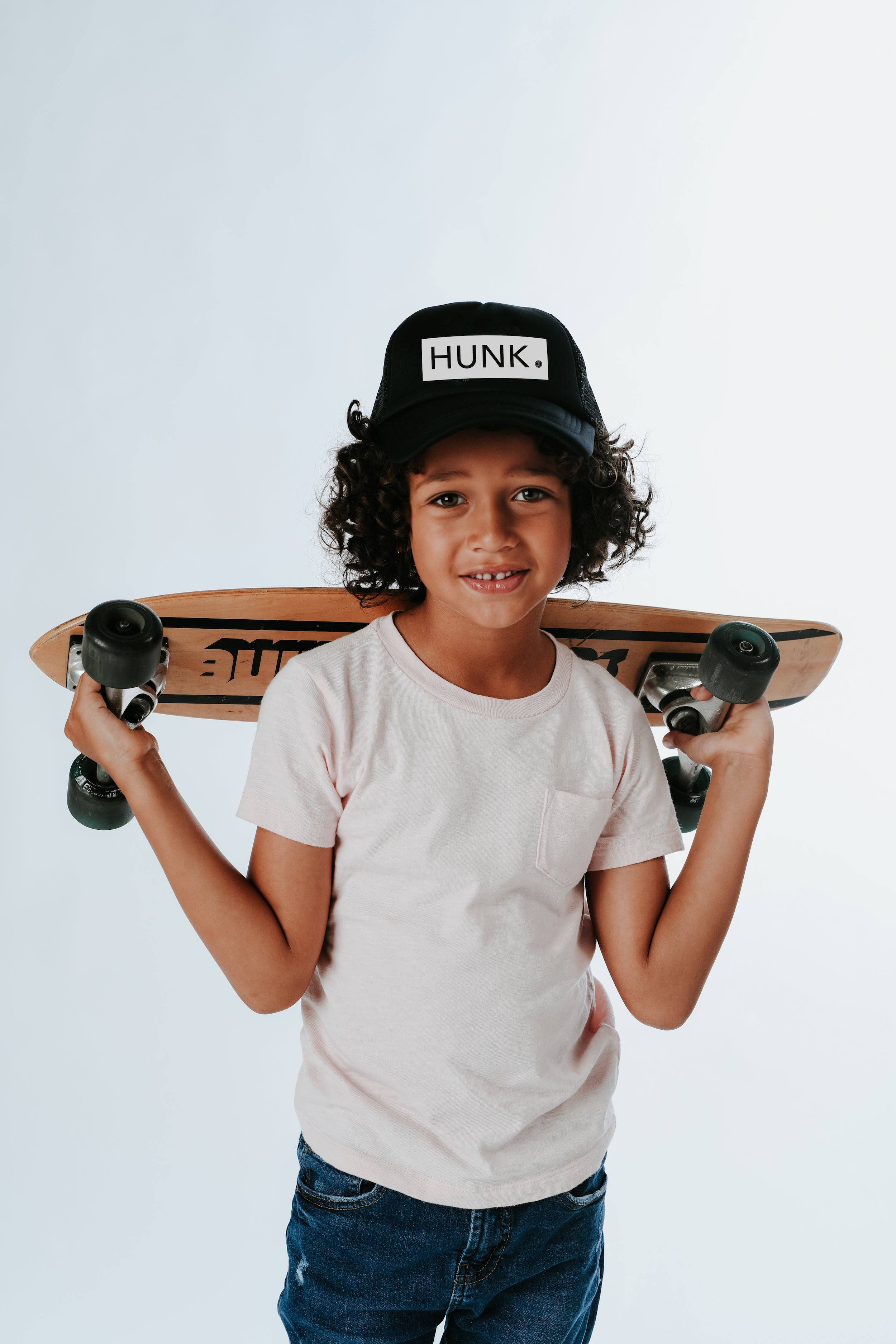 A young child with curly hair smiles while holding a skateboard across their shoulders, donning a Little Hunk Hat from Mama X™ Brand that reads "HUNK." Wearing a light pink T-shirt and blue jeans, they stand against a plain white background. Boy moms will adore this charming scene.