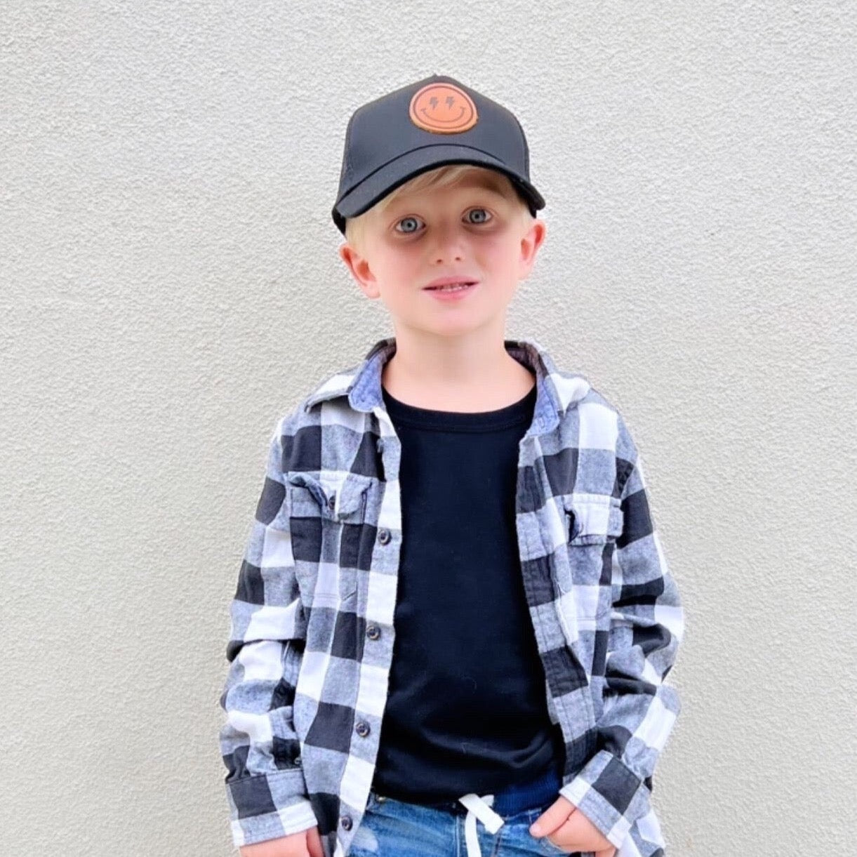 A young boy with blonde hair stands against a light grey wall. He is wearing the Smiley Face Trucker Hat by Mama X™ Brand, featuring a leather smiley face patch, along with a black and white checkered flannel shirt over a black t-shirt, and blue jeans with drawstrings. His hands are casually in his pockets.