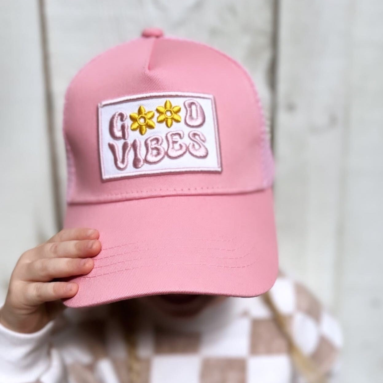 A person wearing the Good Vibes Trucker Hat from Mama X™ Brand, a cute pink cap featuring a patch with "GOOD VIBES" in stylized letters and adorned with two daisy flowers, partially obscures their face. The background is set against a light wooden texture.
