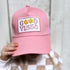 A person wearing the Good Vibes Trucker Hat from Mama X™ Brand, a cute pink cap featuring a patch with "GOOD VIBES" in stylized letters and adorned with two daisy flowers, partially obscures their face. The background is set against a light wooden texture.