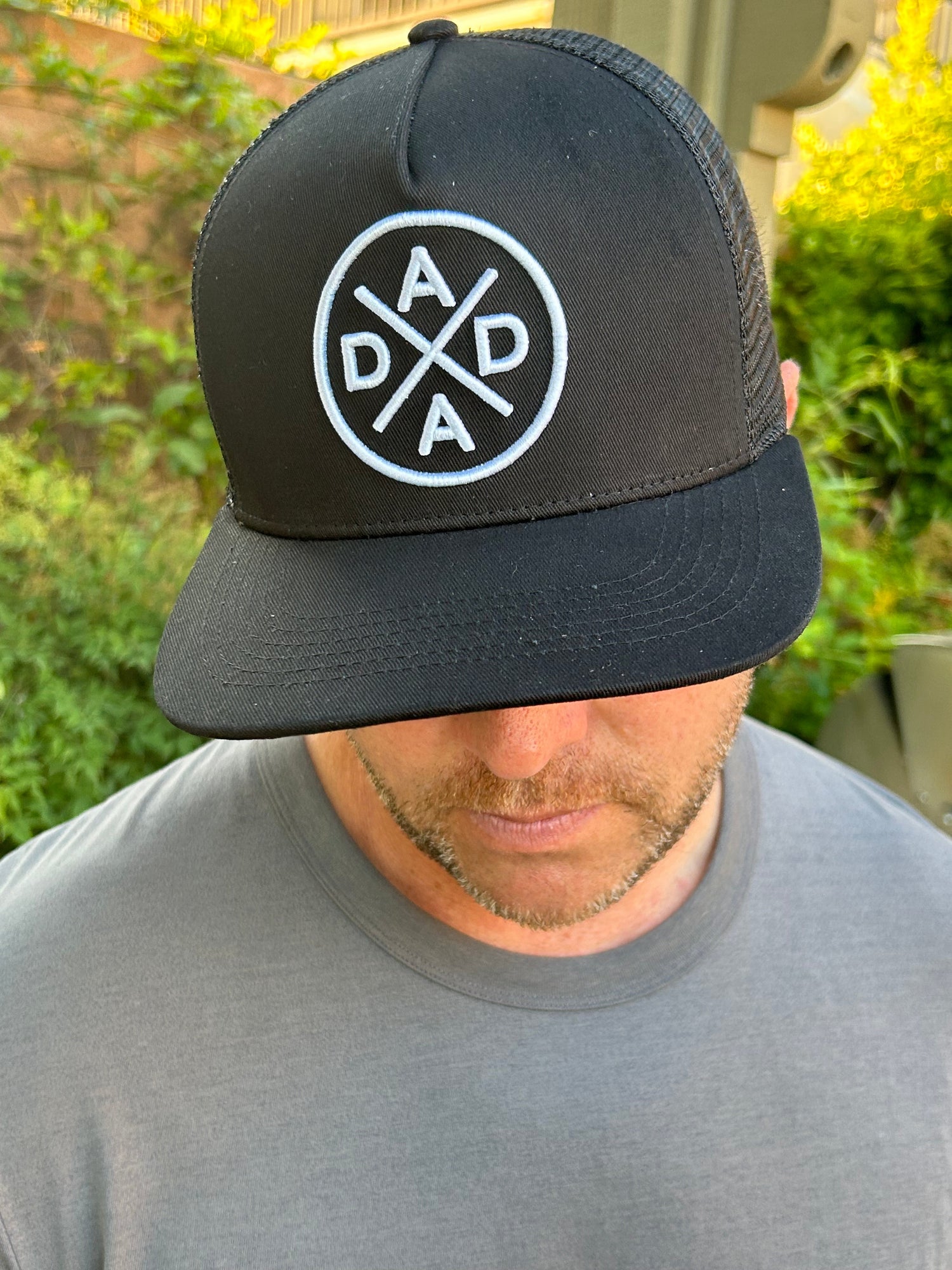 A man wearing the top-selling Dada X™ Premium Trucker Hat-Stitched 3D Logo—a black mesh baseball cap with a circular Dada X™ logo in white letters— is looking downward. He is dressed in a plain grey T-shirt. Green foliage is visible in the blurred background.