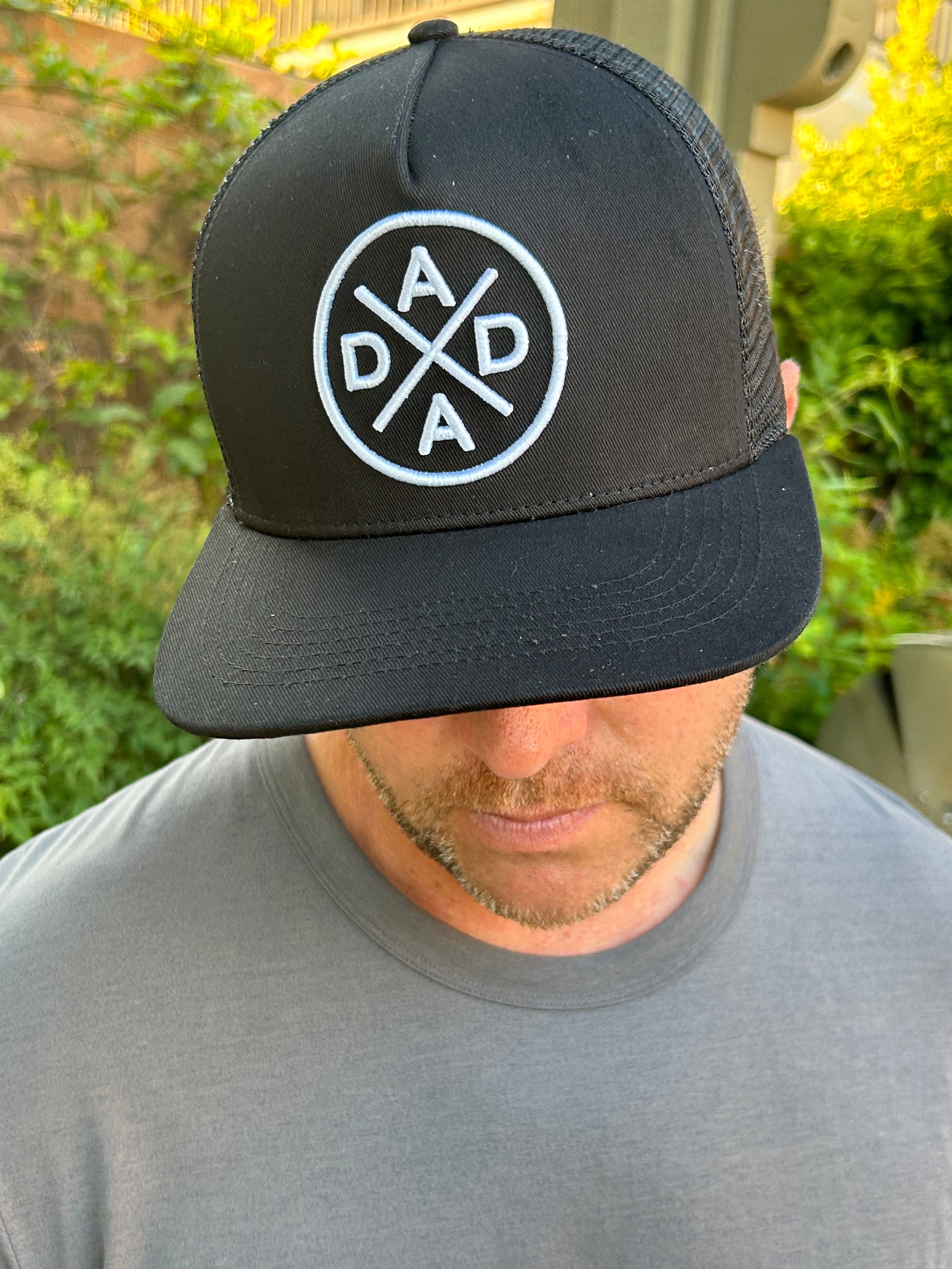 A man wearing the top-selling Dada X™ Premium Trucker Hat-Stitched 3D Logo—a black mesh baseball cap with a circular Dada X™ logo in white letters— is looking downward. He is dressed in a plain grey T-shirt. Green foliage is visible in the blurred background.