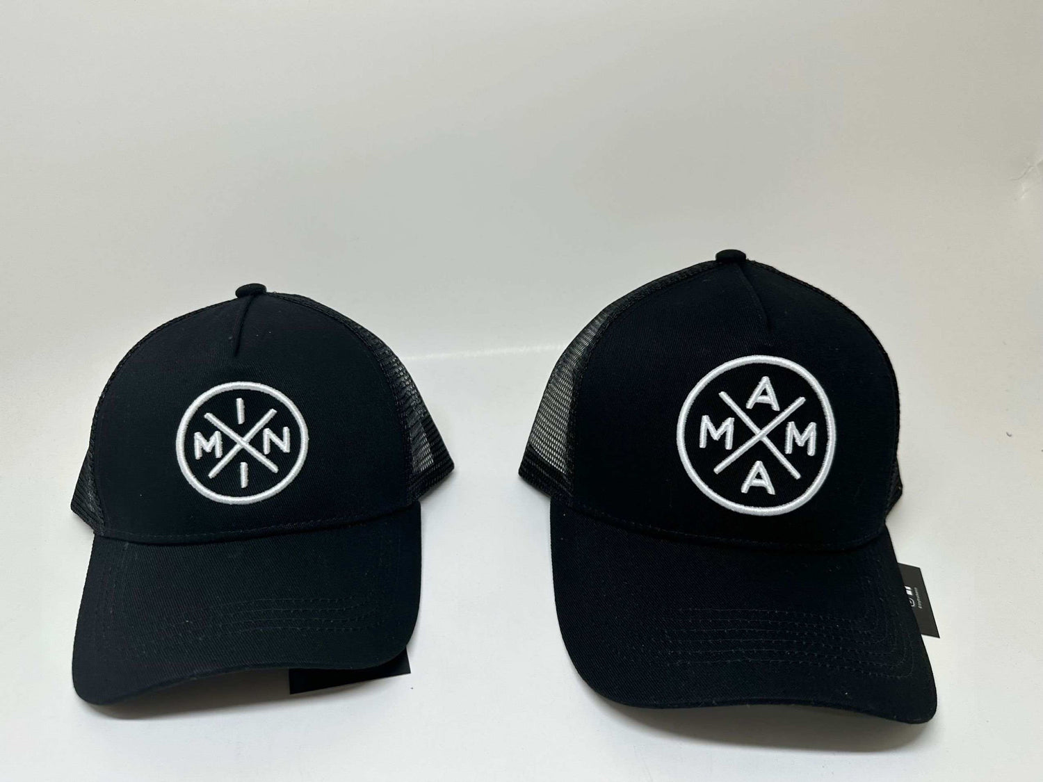 Two black trucker caps are side by side against a white background. The hat on the left is the Mini X Premium Trucker Hat with an embroidered logo featuring intersecting lines and "MIN" and "X" inside. The hat on the right, under the Mama X™ Brand, has a similar design with "AMA" and "X" in the patch.